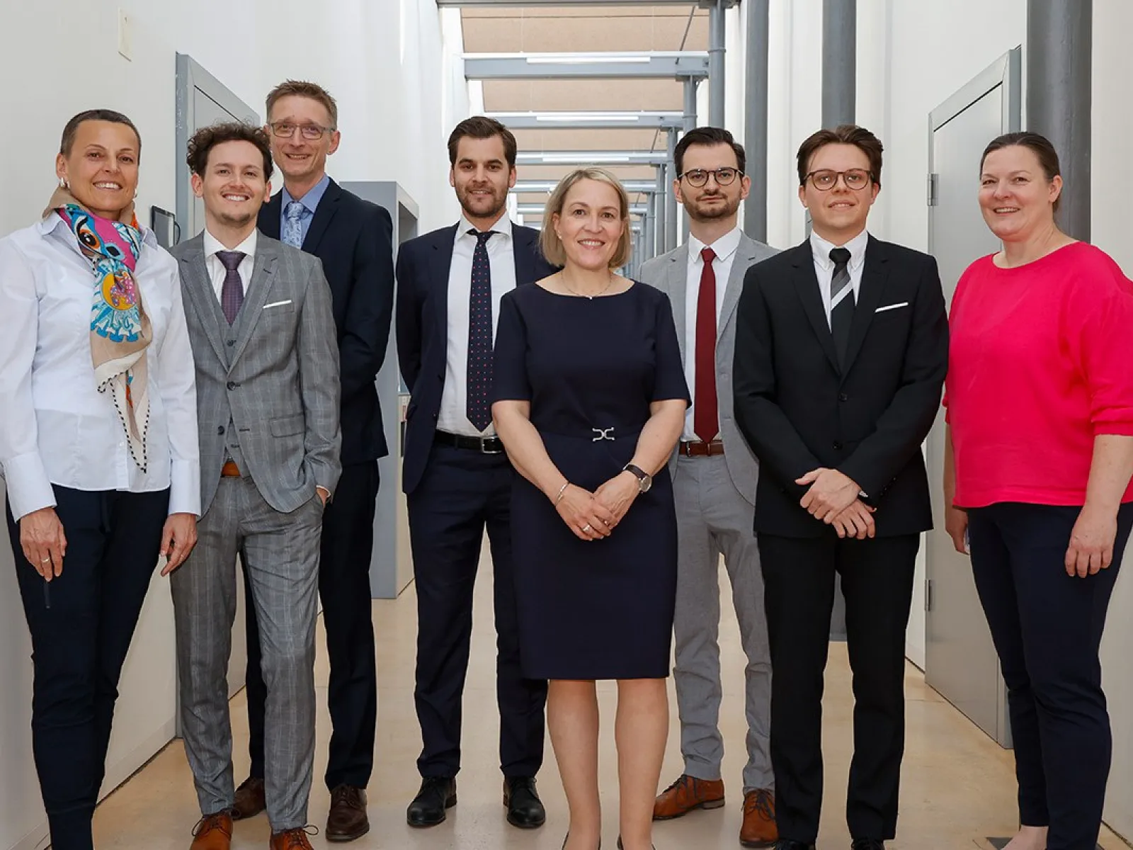 Eine Gruppe von acht Personen steht in einem Flur der Universität Liechtenstein. Sie tragen formelle Kleidung und lächeln in die Kamera. Die Umgebung ist modern und hell gestaltet.