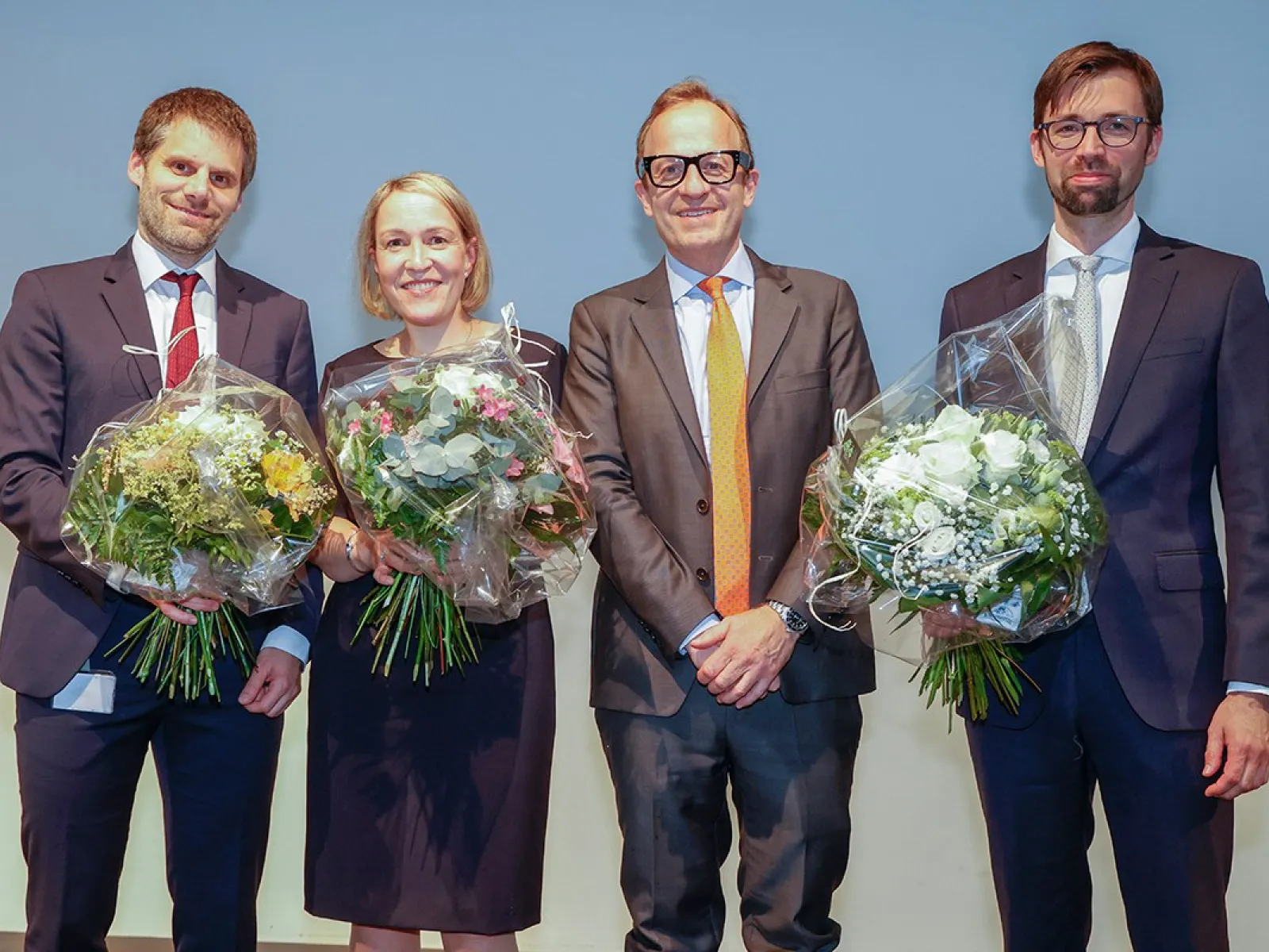 Vier Personen stehen nebeneinander auf einer Bühne. Drei von ihnen halten Blumensträuße in den Händen. Alle tragen formelle Kleidung und lächeln in die Kamera.