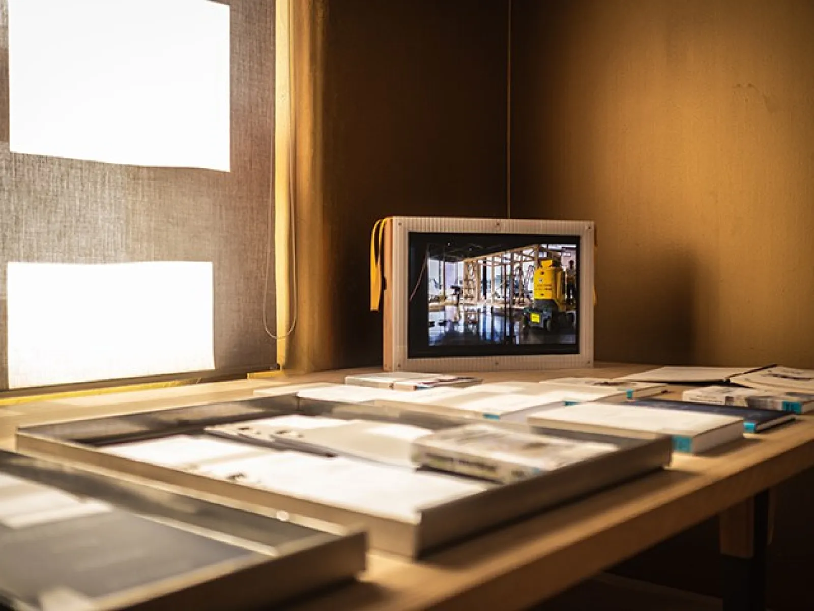 Ein Tisch mit Architekturmodellen und Dokumenten, im Hintergrund ein Bildschirm mit einer Baustellenszene.