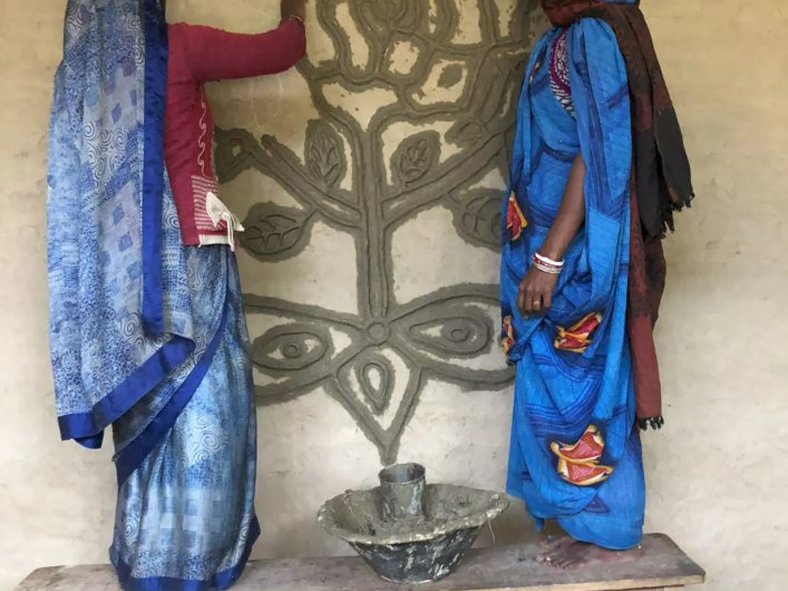 Zwei Frauen in traditionellen indischen Kleidern verzieren eine Wand mit einem kunstvollen Blumenmuster. Sie stehen auf einer Holzbank und arbeiten gemeinsam an der Gestaltung.