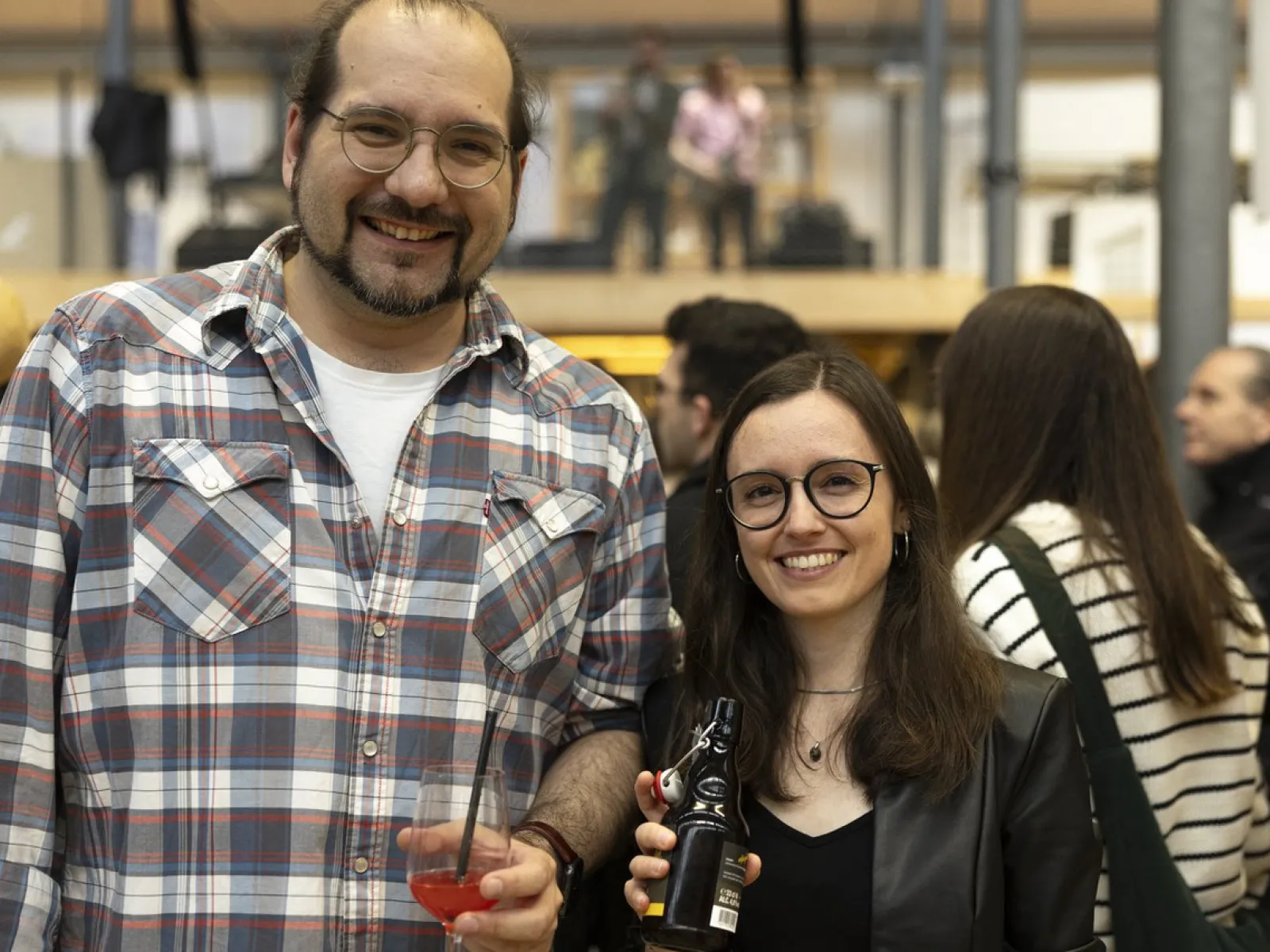 Zwei Personen lächeln in die Kamera bei einer Veranstaltung. Sie halten Getränke in den Händen. Im Hintergrund sind weitere Gäste und eine Bühne zu sehen. Die Atmosphäre wirkt fröhlich und gesellig.