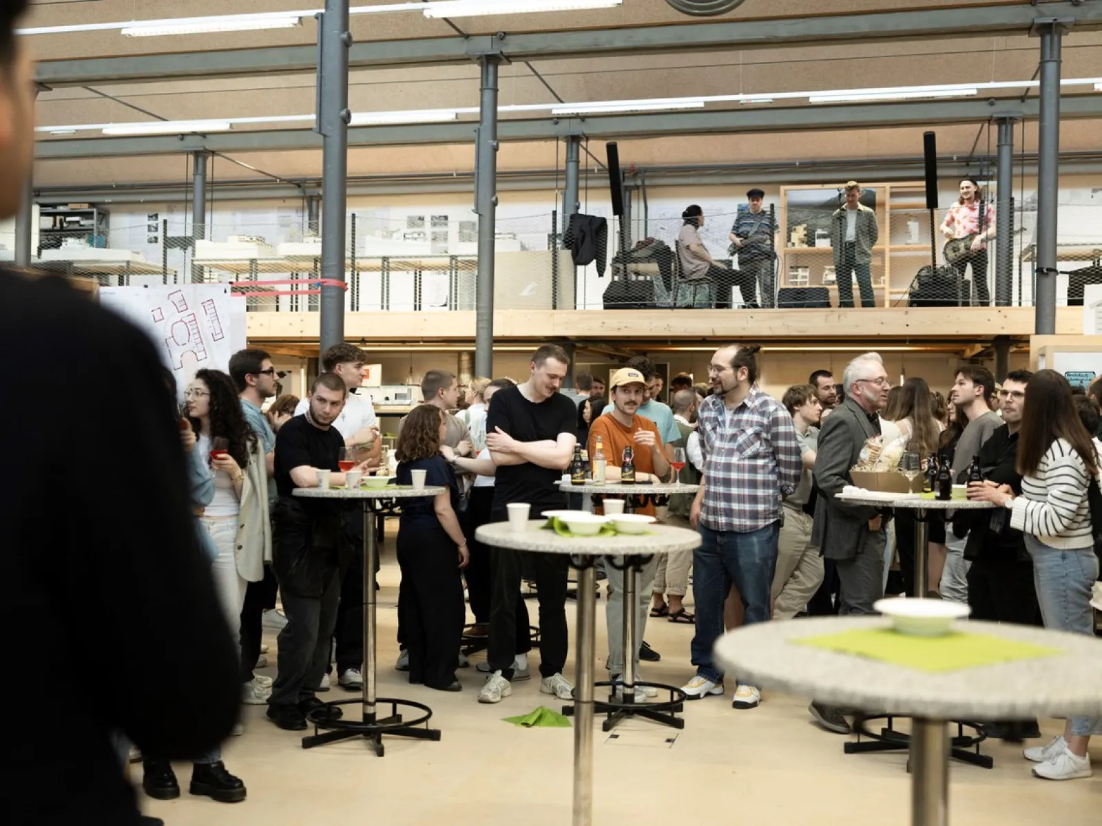 Eine Gruppe von Menschen versammelt sich in einem modernen Innenraum mit hohen Decken und großen Fenstern. Sie stehen um Stehtische, unterhalten sich und genießen die Veranstaltung. Im Hintergrund sind weitere Personen auf einer erhöhten Plattform zu sehen. Die Atmosphäre wirkt lebhaft und gesellig.