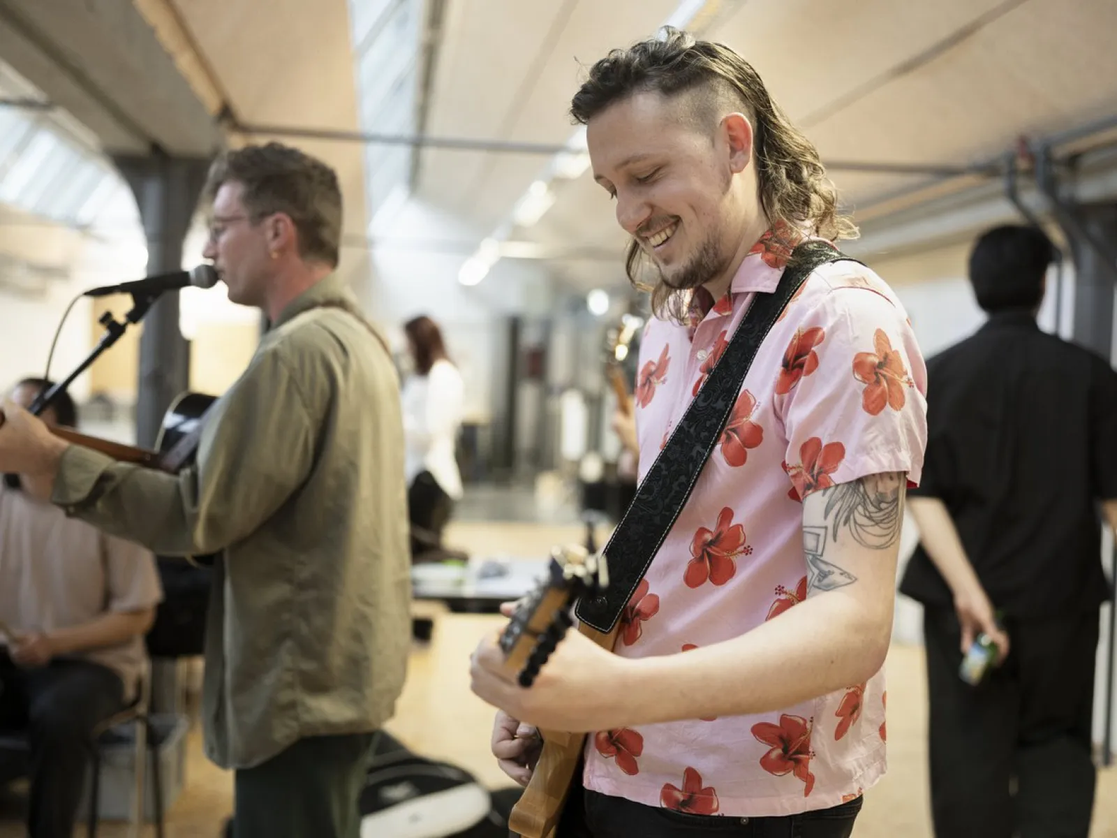 Ein Musiker in einem rosa Hemd mit Blumenmuster spielt Gitarre und lächelt. Im Hintergrund ist ein weiterer Musiker mit Mikrofon zu sehen. Die Szene findet in einem Innenraum statt, möglicherweise während einer Veranstaltung an der Universität Liechtenstein.