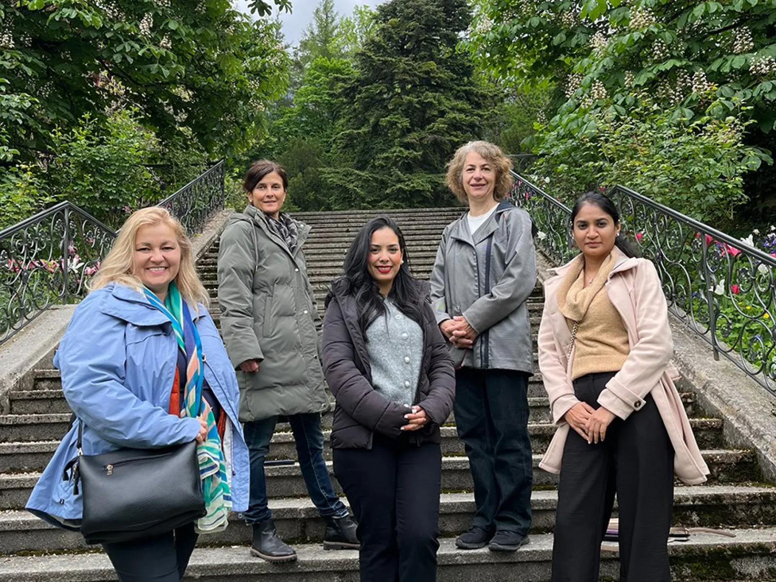 Fünf Personen stehen auf einer Treppe im Freien, umgeben von grünen Bäumen. Sie lächeln in die Kamera.