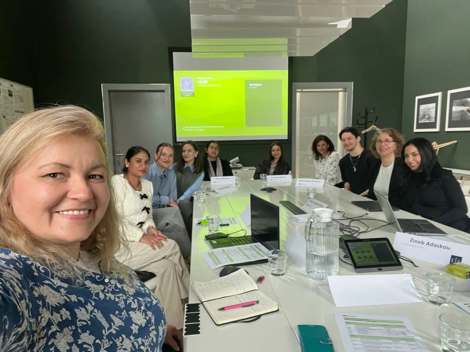 Eine Gruppe von sechs Personen steht lächelnd in einem Raum vor einer Wand mit gerahmten Bildern. Die Personen sind formell gekleidet und blicken in die Kamera.