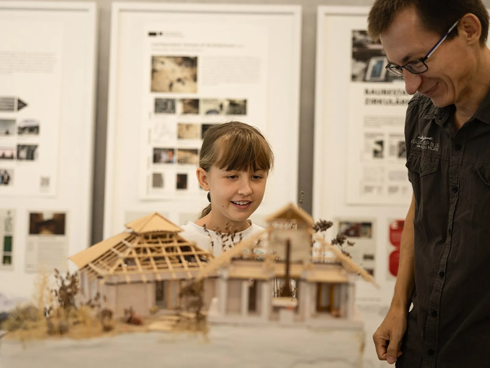 Ein Kind und ein Erwachsener betrachten ein detailliertes Architekturmodell eines Gebäudes. Im Hintergrund sind Informationsplakate zu sehen.