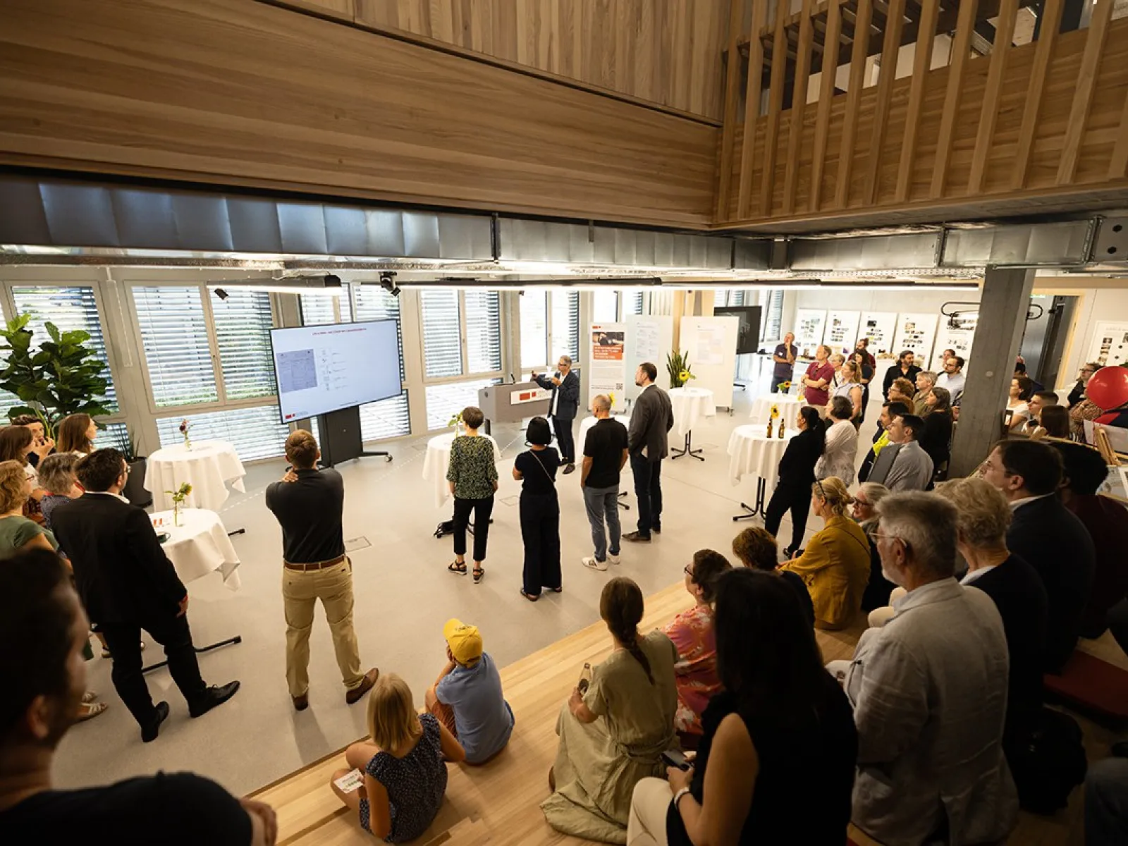Eine Gruppe von Menschen versammelt sich in einem modernen, hellen Raum mit großen Fenstern. Im Vordergrund sitzen einige Personen auf Holzstufen. Im Hintergrund hält eine Person eine Präsentation vor einem Bildschirm. Der Raum ist mit Pflanzen und Stehtischen dekoriert.