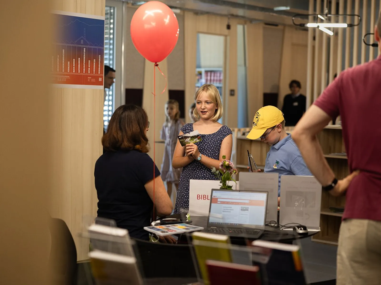 Eine Gruppe von Menschen steht an einem Informationsstand in einem modernen Gebäude. Eine Frau sitzt hinter dem Tisch, während ein Mädchen mit einem roten Ballon in der Hand lächelt. Ein Junge mit gelber Kappe betrachtet die ausgestellten Materialien. Im Hintergrund sind Regale und weitere Personen zu sehen.