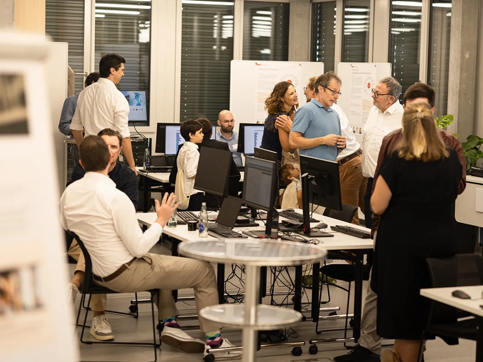 Eine Gruppe von Menschen unterhält sich in einem modernen Raum mit Computern und Informationspostern. Einige sitzen, während andere stehen und diskutieren. Die Atmosphäre wirkt lebhaft und interessiert, passend zu einem Tag der offenen Tür an der Universität.