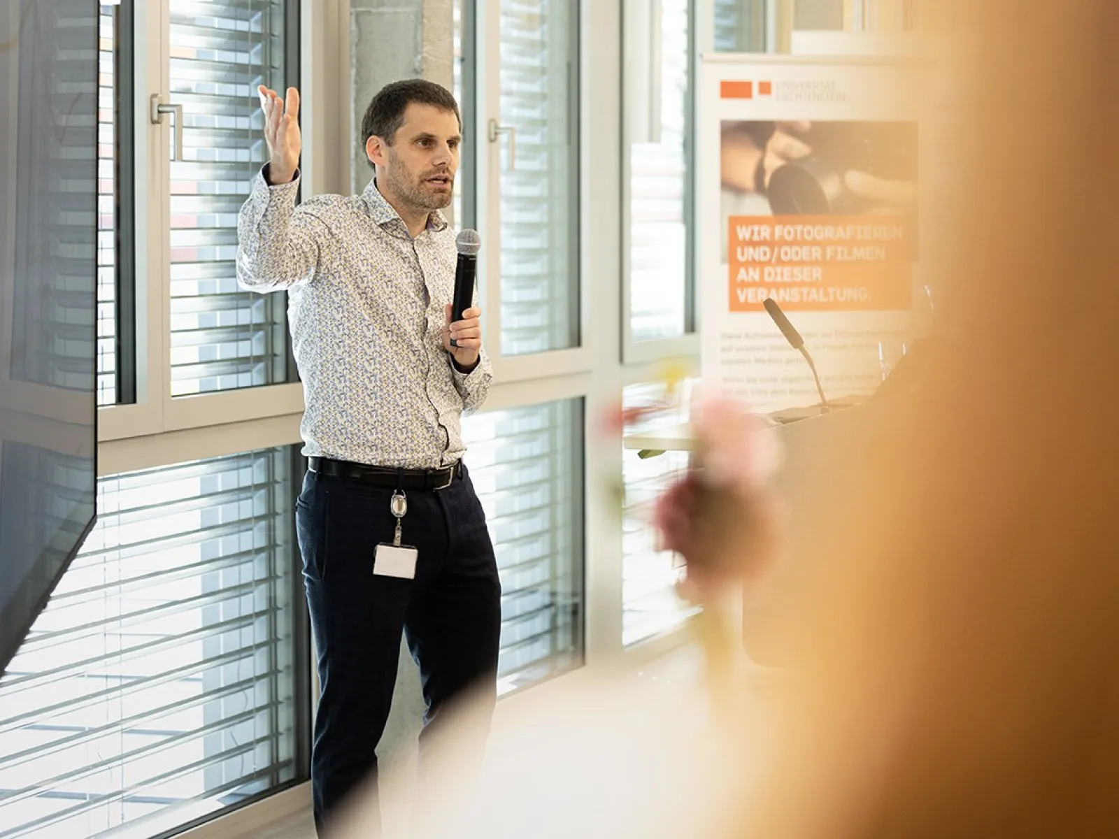 Eine Person hält eine Präsentation in einem modernen Raum mit großen Fenstern. Sie spricht in ein Mikrofon und gestikuliert mit der Hand. Im Hintergrund ist ein Plakat mit Informationen sichtbar.