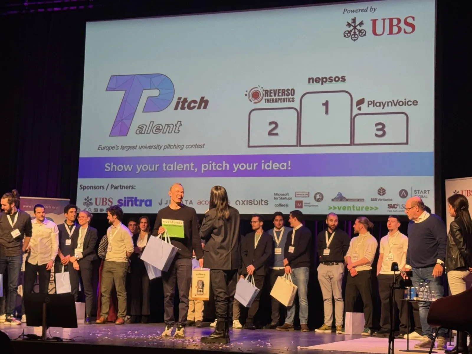 Eine Gruppe von Menschen steht auf einer Bühne beim Talent Pitch Switzerland im Volkshaus Zürich. Im Hintergrund ist eine Leinwand mit den Logos der Gewinner-Start-ups Reverso Therapeutics, Nepsos und PlaynVoice zu sehen. Die Veranstaltung wird von UBS gesponsert.