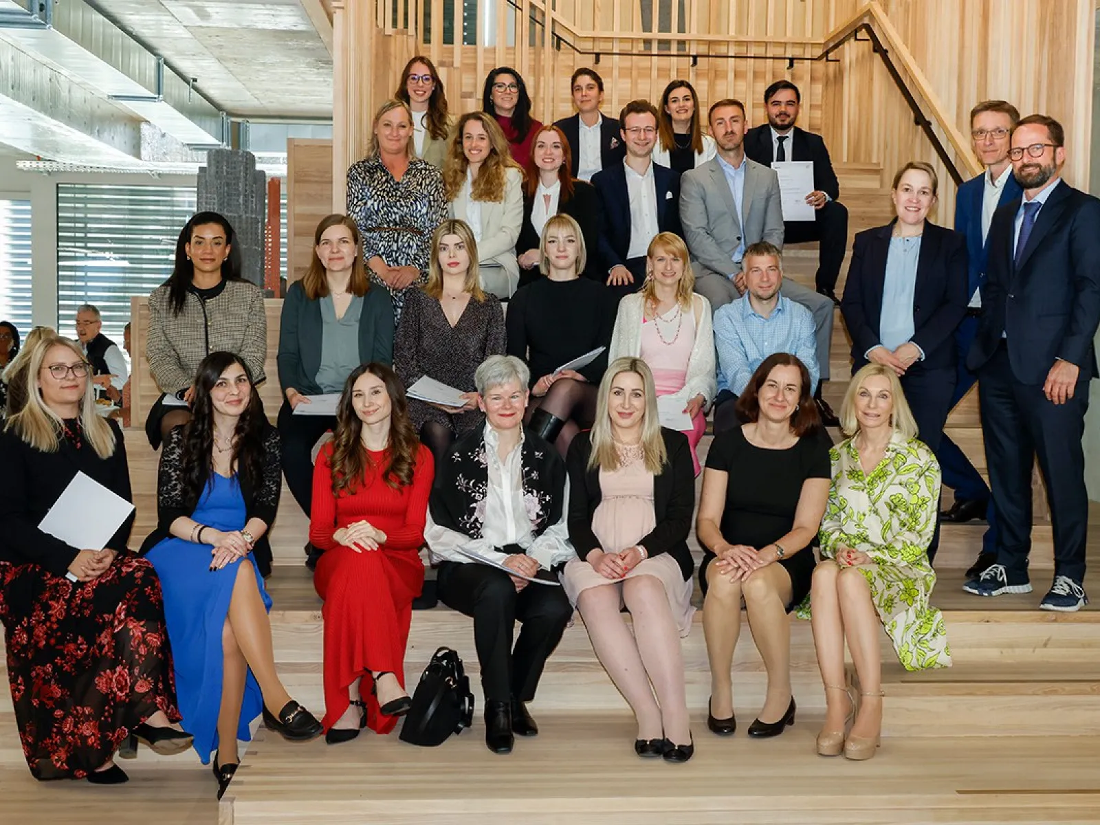 Gruppenfoto von Absolventinnen und Absolventen des Zertifikatsstudiengangs Treuhandwesen der Universität Liechtenstein, die auf einer Treppe sitzen und stehen. Sie halten Zertifikate in den Händen und lächeln in die Kamera. Im Hintergrund ist ein moderner Innenraum zu sehen.