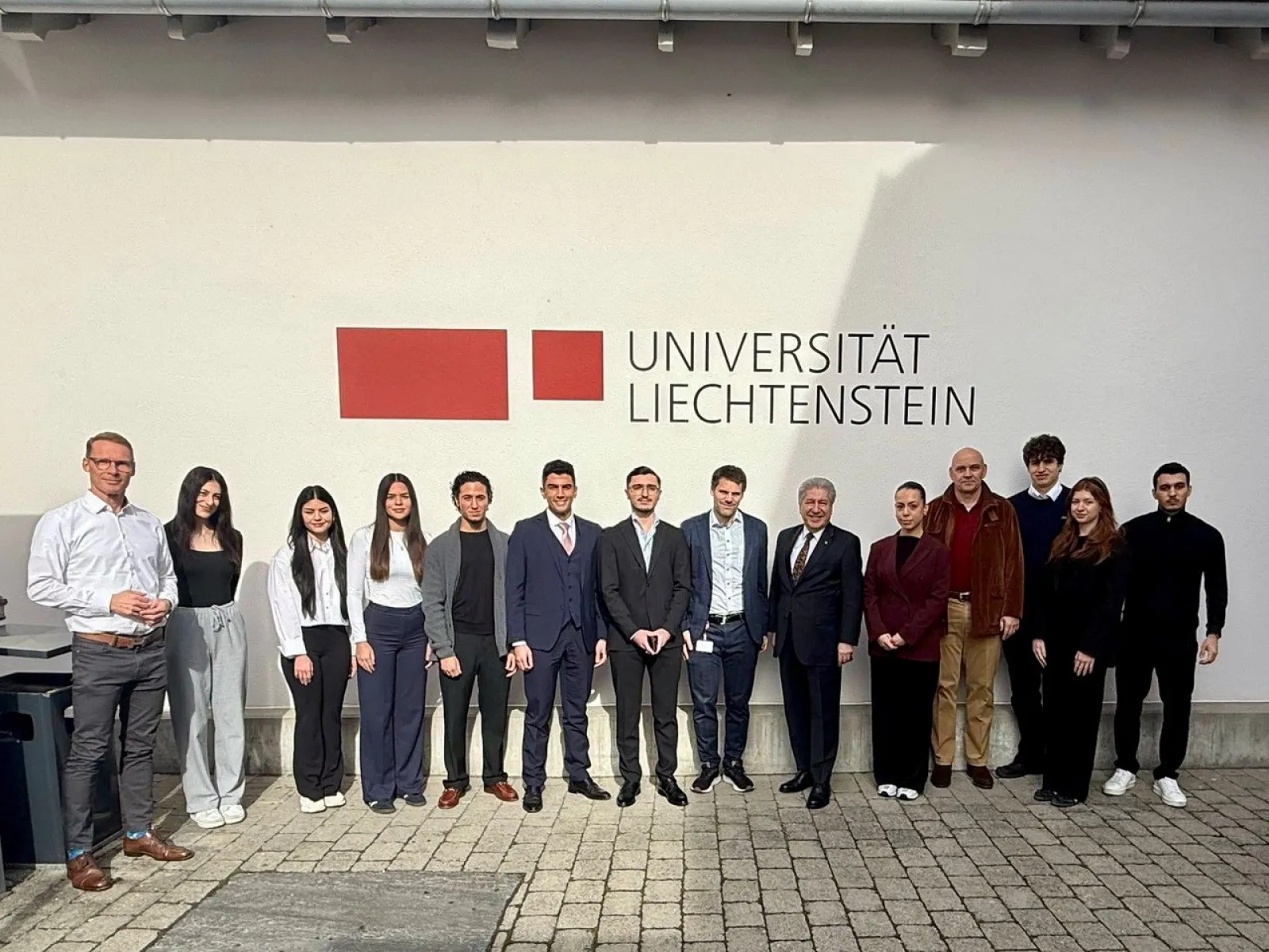 Eine Gruppe von Menschen steht vor einer Wand mit dem Logo der Universität Liechtenstein. Sie sind formell gekleidet und lächeln in die Kamera.