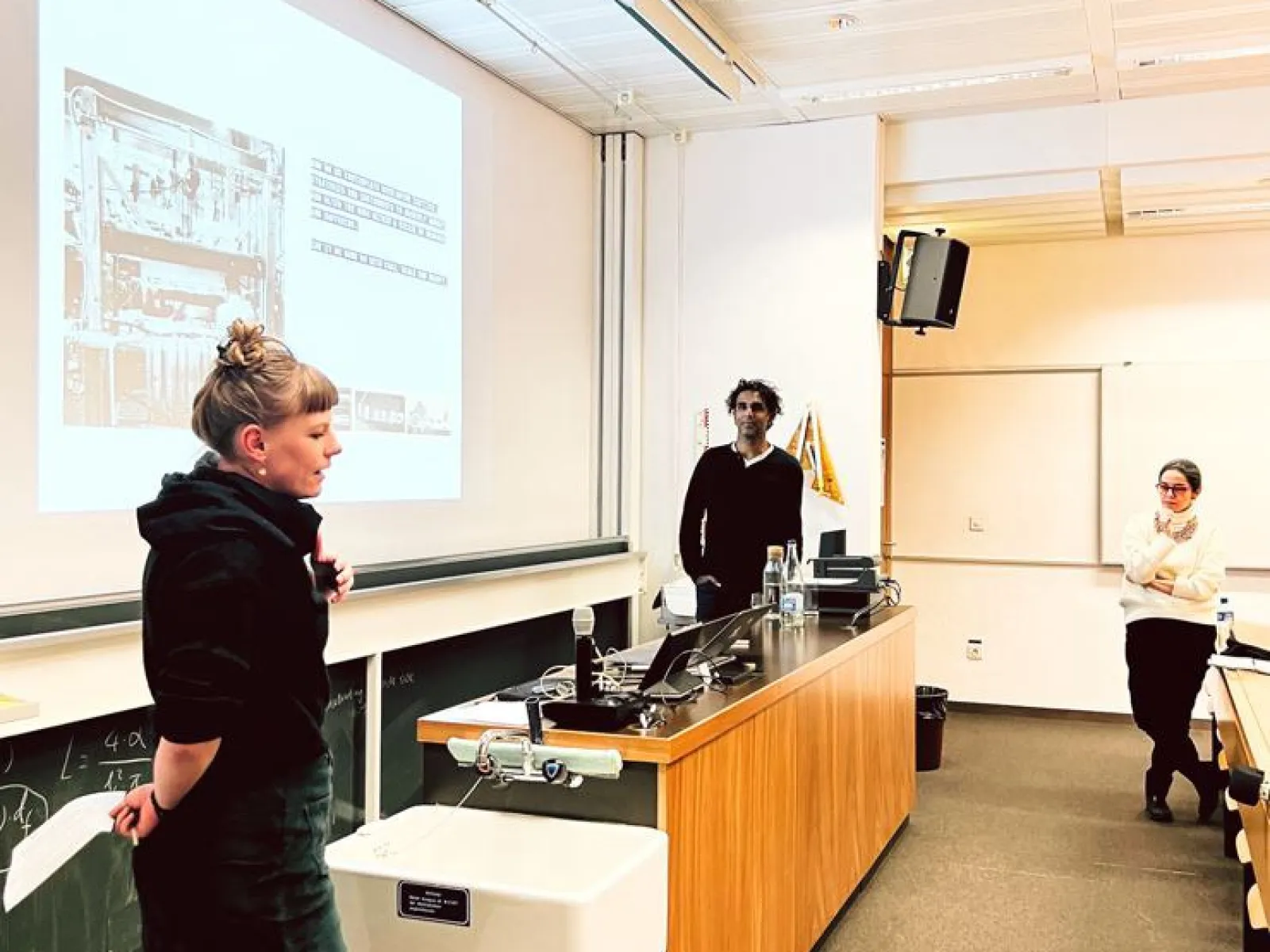 Eine Präsentation in einem Universitätsseminarraum: Drei Personen stehen vor einer Leinwand, auf der eine Folie projiziert wird. Eine Person spricht, während die anderen zuhören. Der Raum ist mit Tischen und einer Tafel ausgestattet.