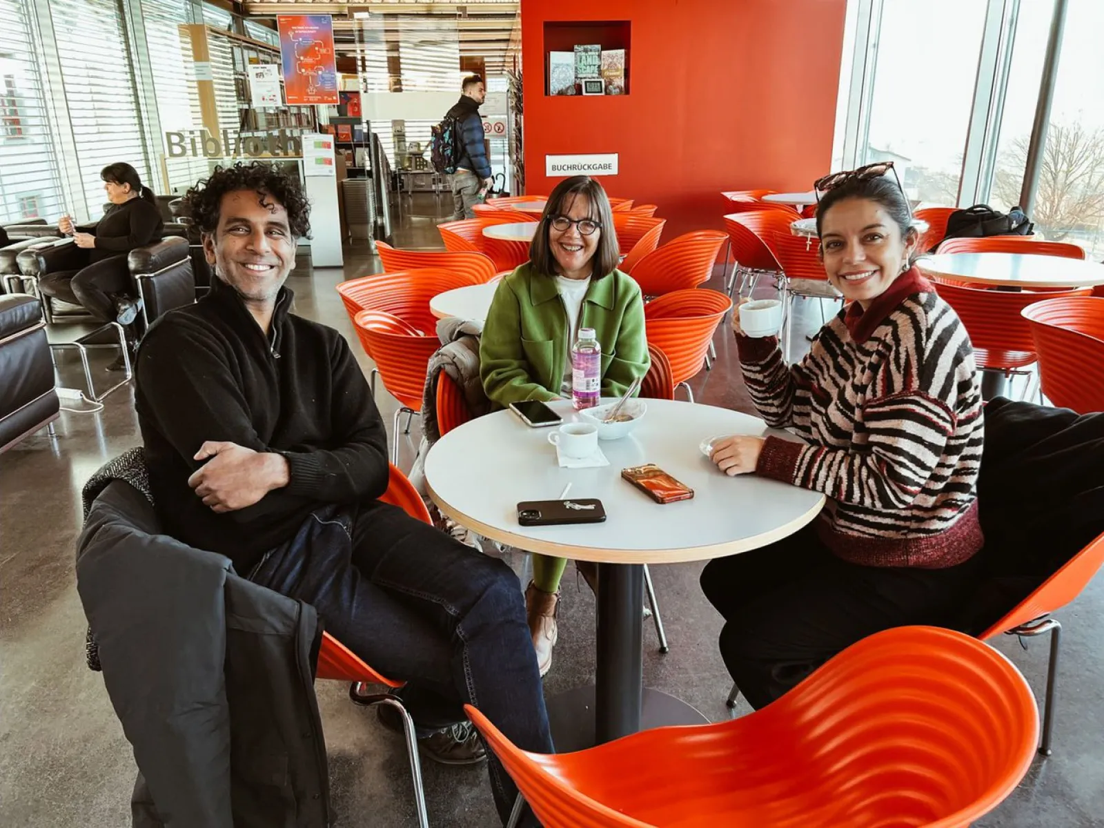 Drei Personen sitzen lächelnd an einem Tisch in einem modernen Café mit roten Stühlen. Sie halten Getränke in der Hand und wirken entspannt. Im Hintergrund sind weitere Gäste und eine Bücherwand zu sehen.