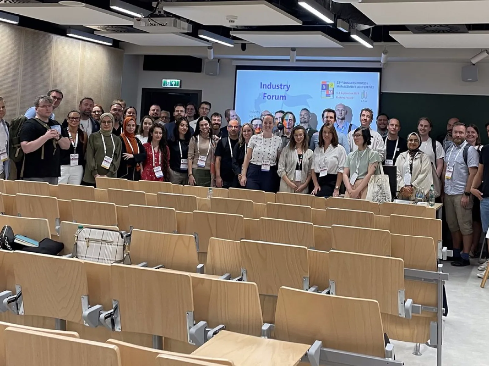 Eine Gruppe von Menschen steht in einem Hörsaal vor einer Präsentationsleinwand. Die Leinwand zeigt den Titel "Industry Forum". Die Personen lächeln in die Kamera. Im Vordergrund sind leere Sitzreihen zu sehen.