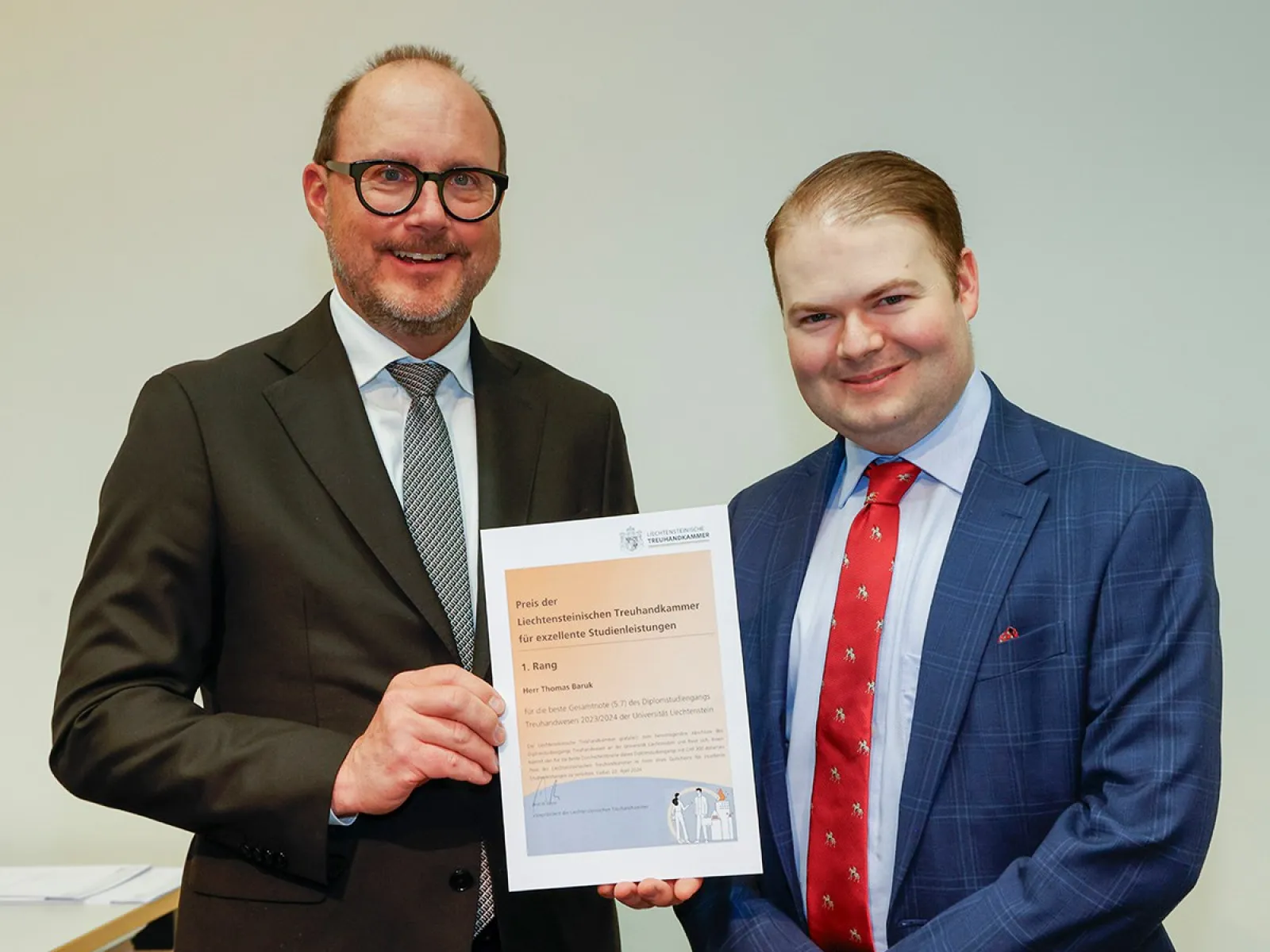 Zwei Männer in Anzügen halten gemeinsam ein Zertifikat in die Kamera. Der Hintergrund ist neutral. Sie lächeln und stehen in einem Raum, der für eine feierliche Veranstaltung geeignet wirkt.