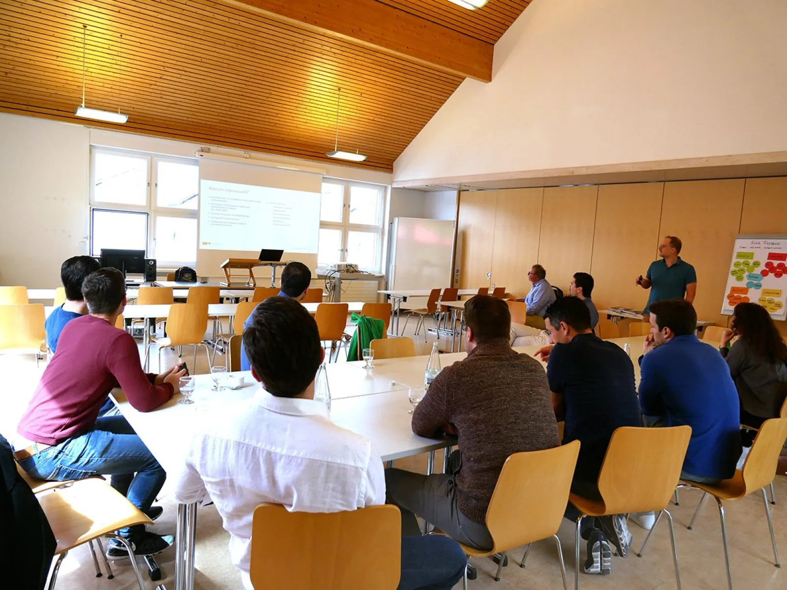 Eine Gruppe von Menschen sitzt in einem Seminarraum der Universität Liechtenstein und hört einer Präsentation zu. Ein Mann steht vorne und spricht, während eine Präsentation auf eine Leinwand projiziert wird. Die Teilnehmer sitzen an Tischen und sind aufmerksam.