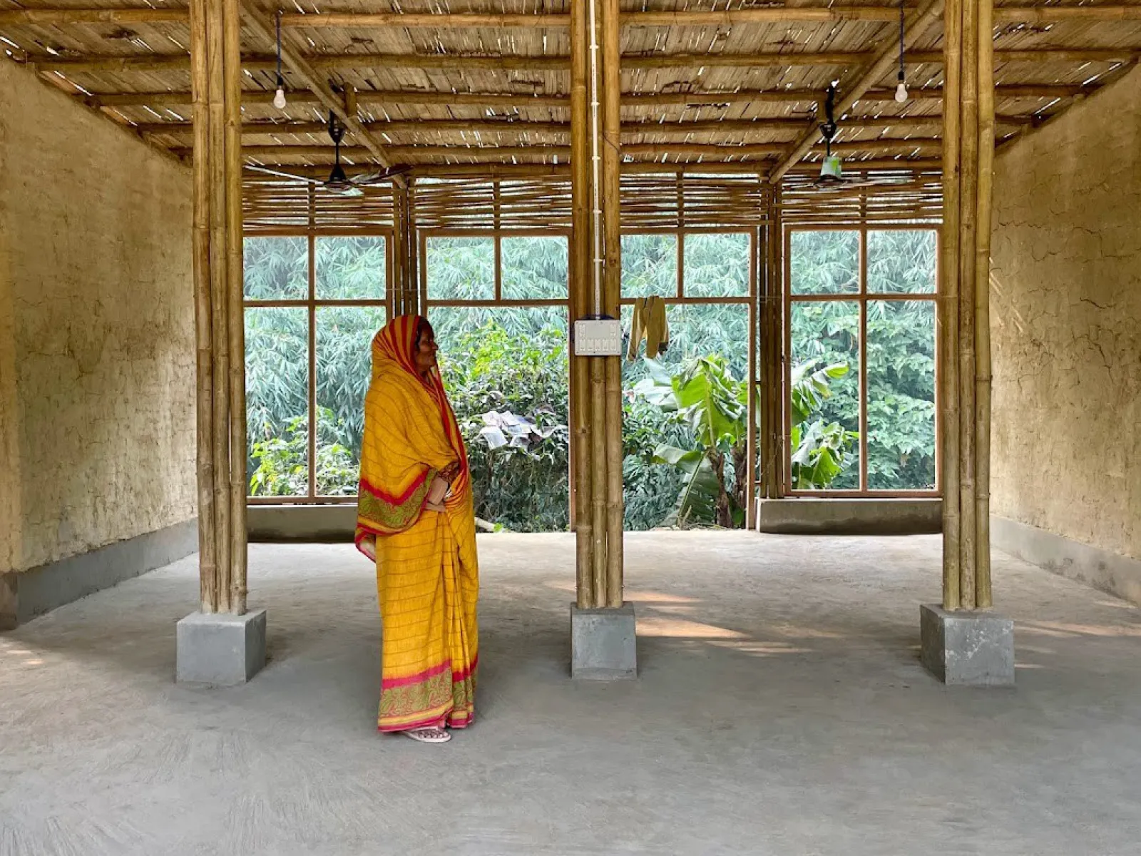 Eine Frau in traditioneller indischer Kleidung steht in einem offenen Raum mit Lehmwänden und Bambussäulen. Große Fenster im Hintergrund bieten einen Blick auf üppiges Grün.
