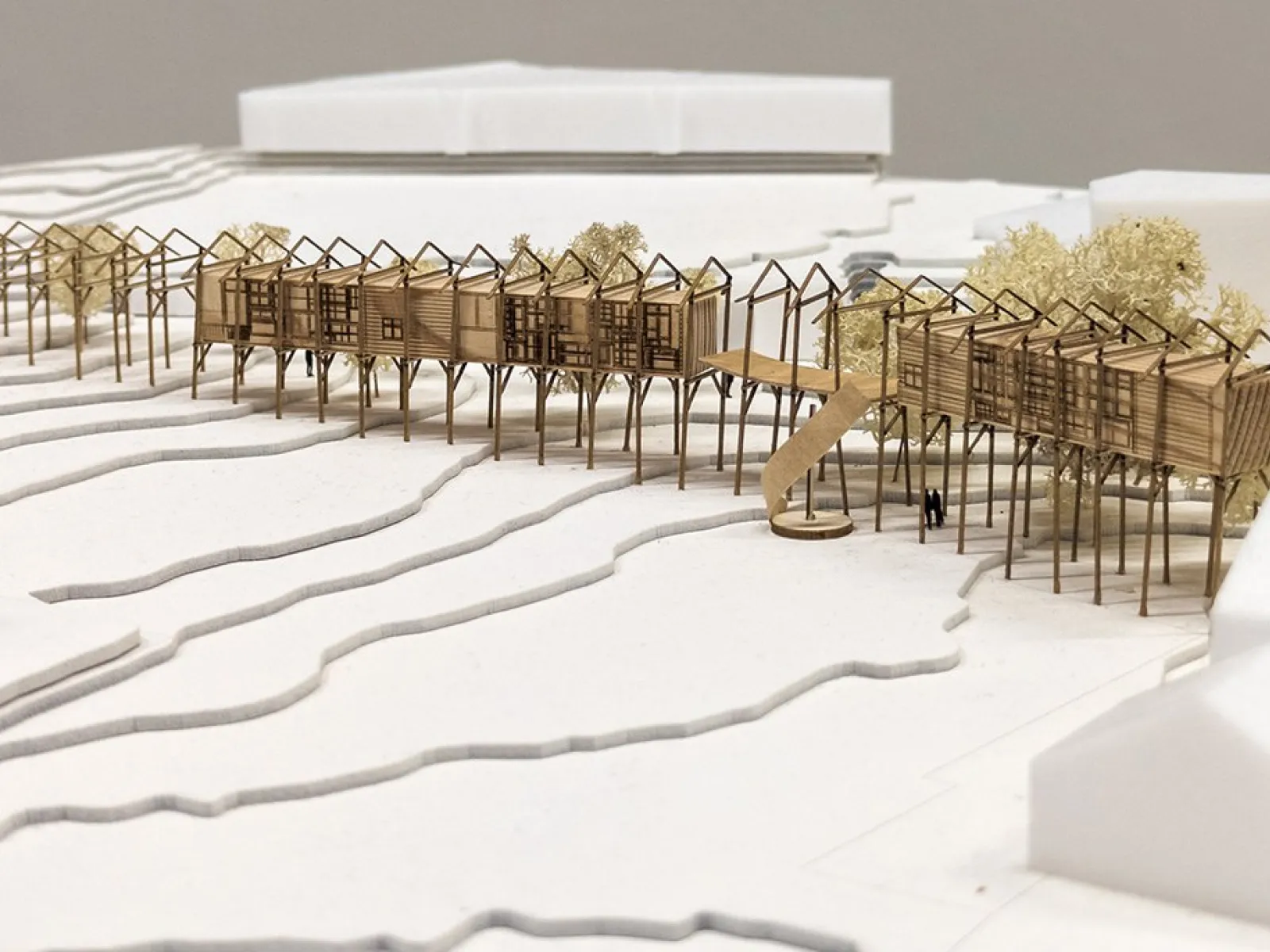Architekturmodell eines linearen Gebäudekomplexes auf Stelzen, eingebettet in eine Landschaft mit Höhenlinien. Das Modell zeigt eine innovative Wiederverwendung von Bauteilen eines alten Strickbaus, umgeben von stilisierten Bäumen und Gebäuden.