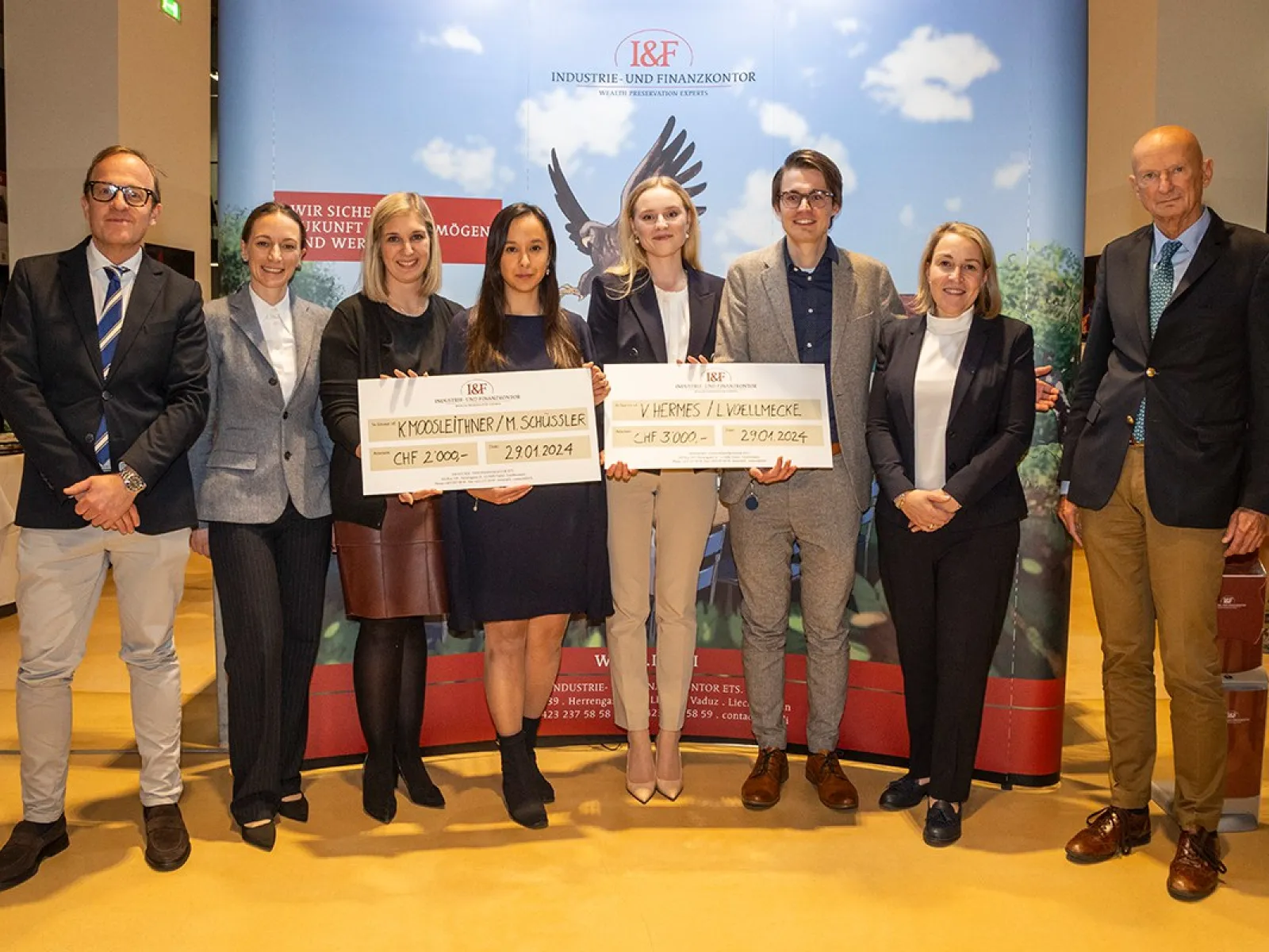 Gruppe von acht Personen posiert mit zwei großen Schecks bei der Verleihung des I&F Family Wealth Preservation Awards an der Universität Liechtenstein. Die Studierenden wurden für ihre herausragenden Strukturierungsvorschläge ausgezeichnet. Im Hintergrund ist ein Banner des Industrie- und Finanzkontors zu sehen.