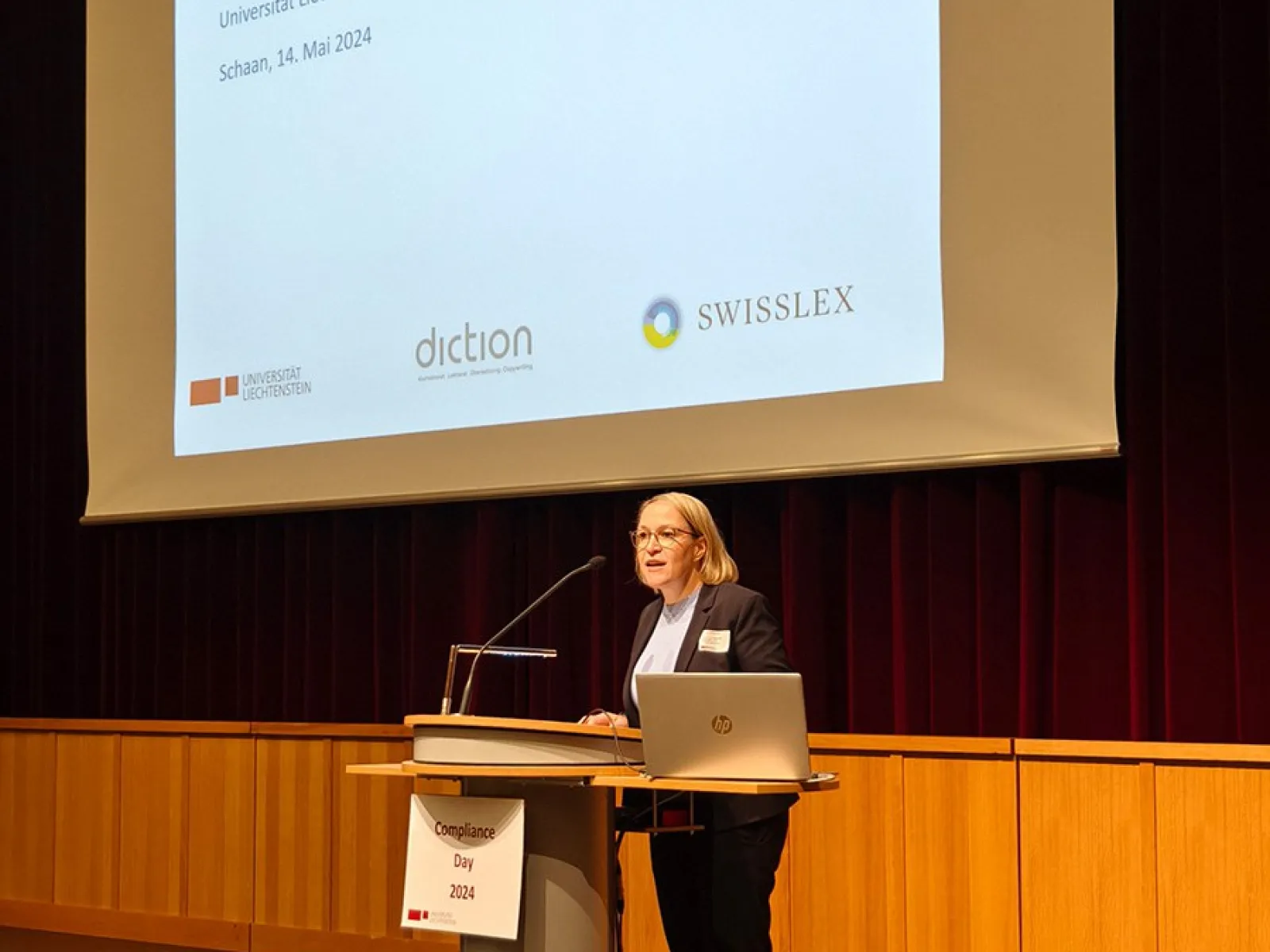 Eine Frau steht an einem Rednerpult vor einer Leinwand mit der Aufschrift „Herzlich willkommen! Compliance-Day 2024, Universität Liechtenstein, Schaan, 14. Mai 2024“. Im Hintergrund sind Logos von Sponsoren zu sehen.