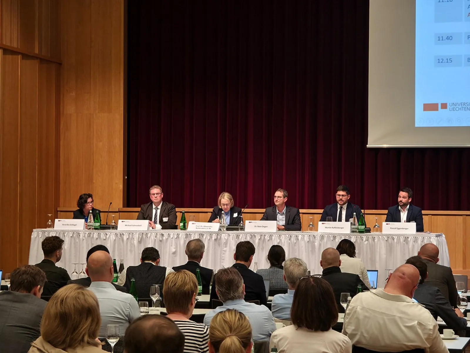 Ein Podium mit sechs Personen sitzt vor einem Publikum in einem Konferenzsaal. Auf dem Tisch stehen Namensschilder und Wasserflaschen. Im Hintergrund ist ein roter Vorhang und ein Teil einer Präsentationsleinwand zu sehen.