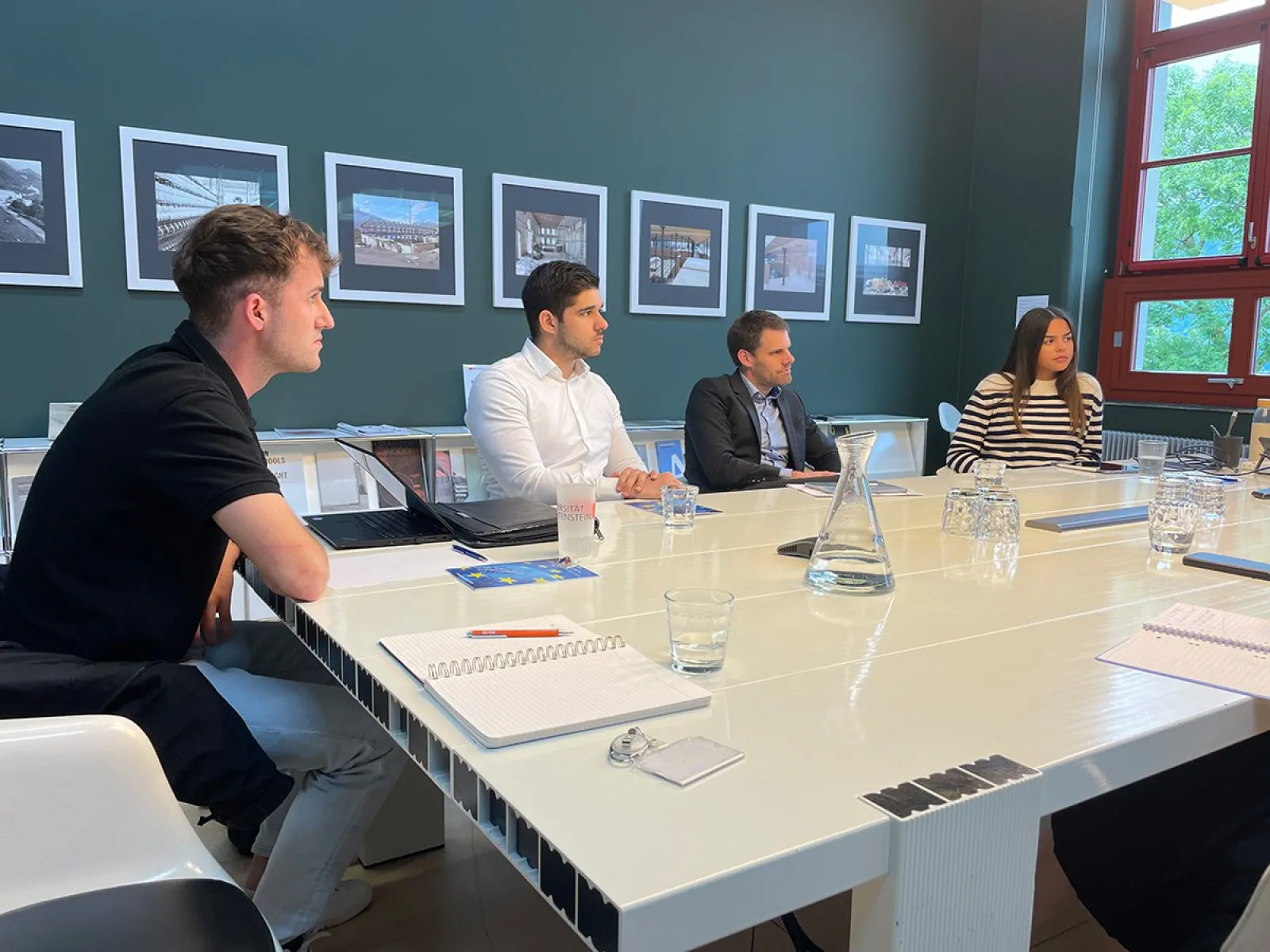 Eine Gruppe von vier Personen sitzt an einem weißen Konferenztisch in einem Raum mit grüner Wand und mehreren gerahmten Bildern. Auf dem Tisch liegen Notizbücher, Stifte und Gläser. Die Personen wirken konzentriert und diskutieren möglicherweise ein Thema. Im Hintergrund sind große Fenster zu sehen, die Tageslicht hereinlassen.