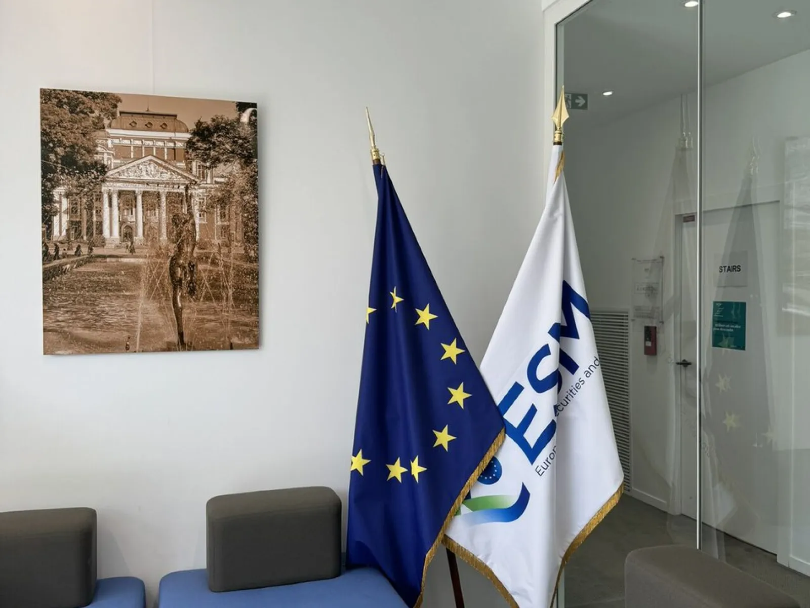 Ein moderner Warteraum mit blauen Sitzmöbeln, zwei Flaggen – eine der Europäischen Union und eine mit dem Logo des Europarats – und einem gerahmten Bild eines historischen Gebäudes an der Wand.