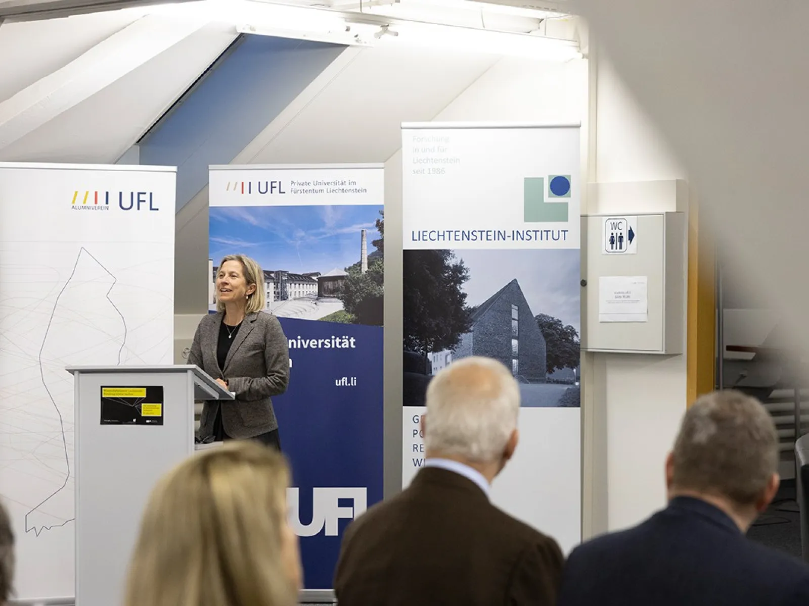 Eine Frau steht an einem Rednerpult vor Bannern der Universität Liechtenstein, der UFL und des Liechtenstein-Instituts. Im Vordergrund sind unscharf einige Zuhörer zu sehen.
