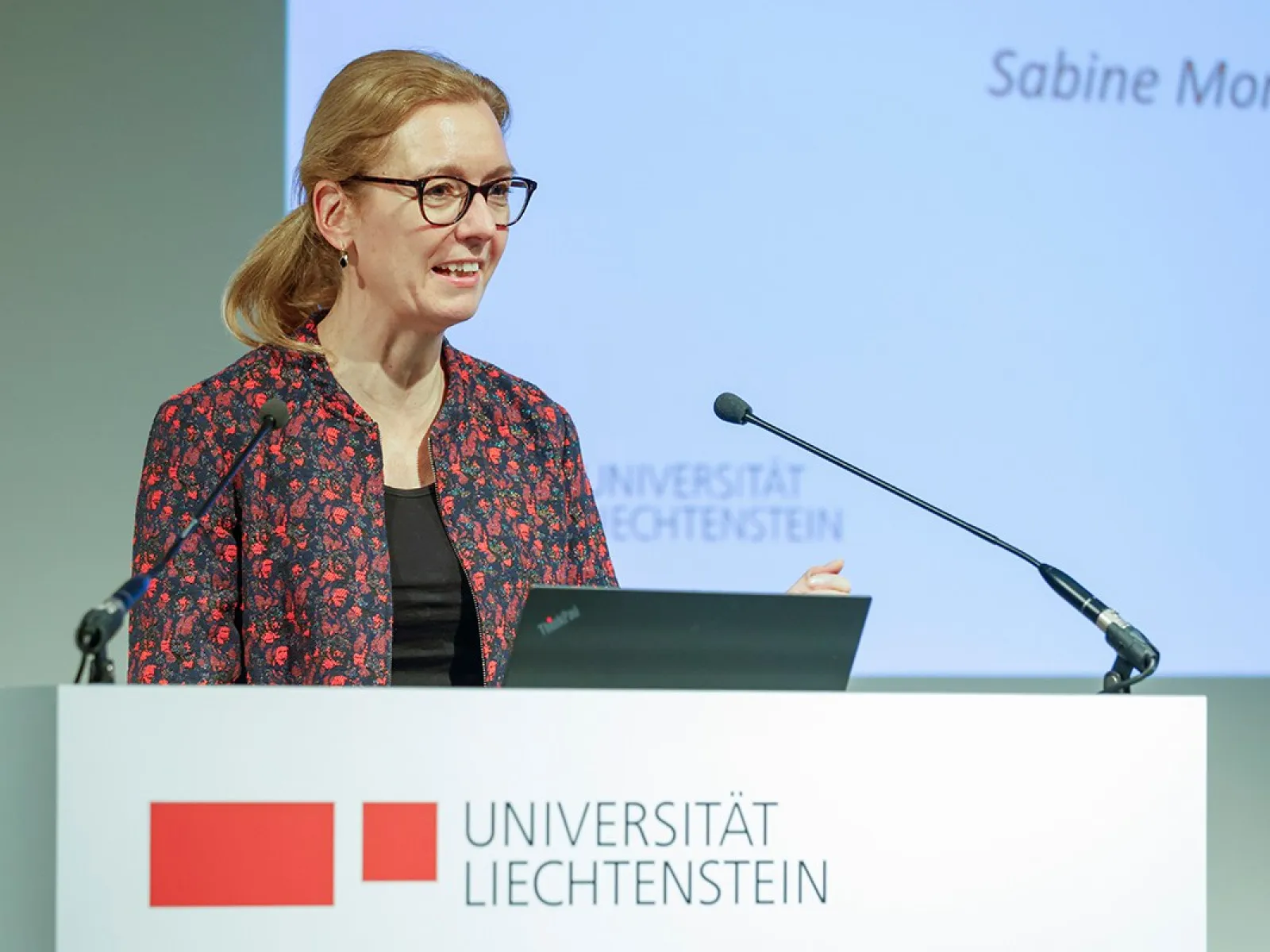 Eine Person hält eine Rede an einem Rednerpult mit dem Logo der Universität Liechtenstein. Im Hintergrund ist eine Präsentation zu sehen.