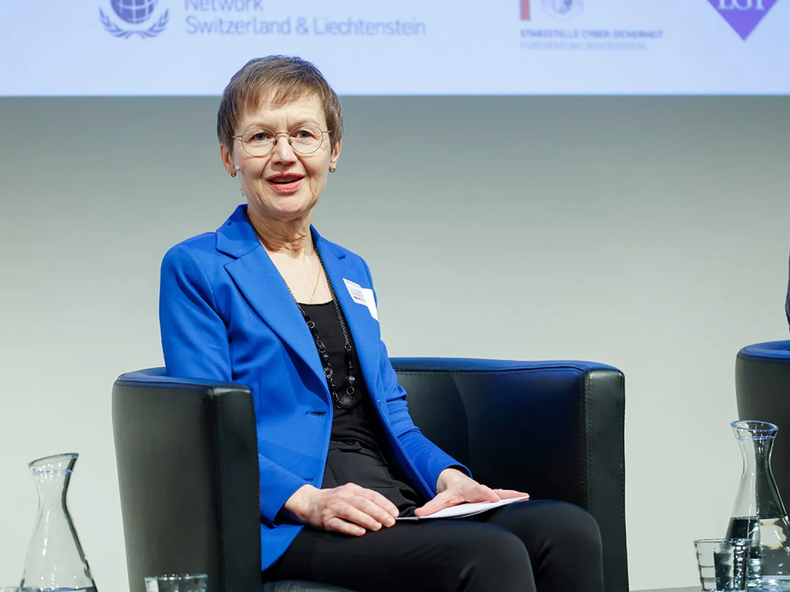 Eine Person in einem blauen Blazer sitzt auf einem Podium und spricht bei einer Veranstaltung an der Universität Liechtenstein. Im Hintergrund sind Logos von Partnerorganisationen zu sehen.
