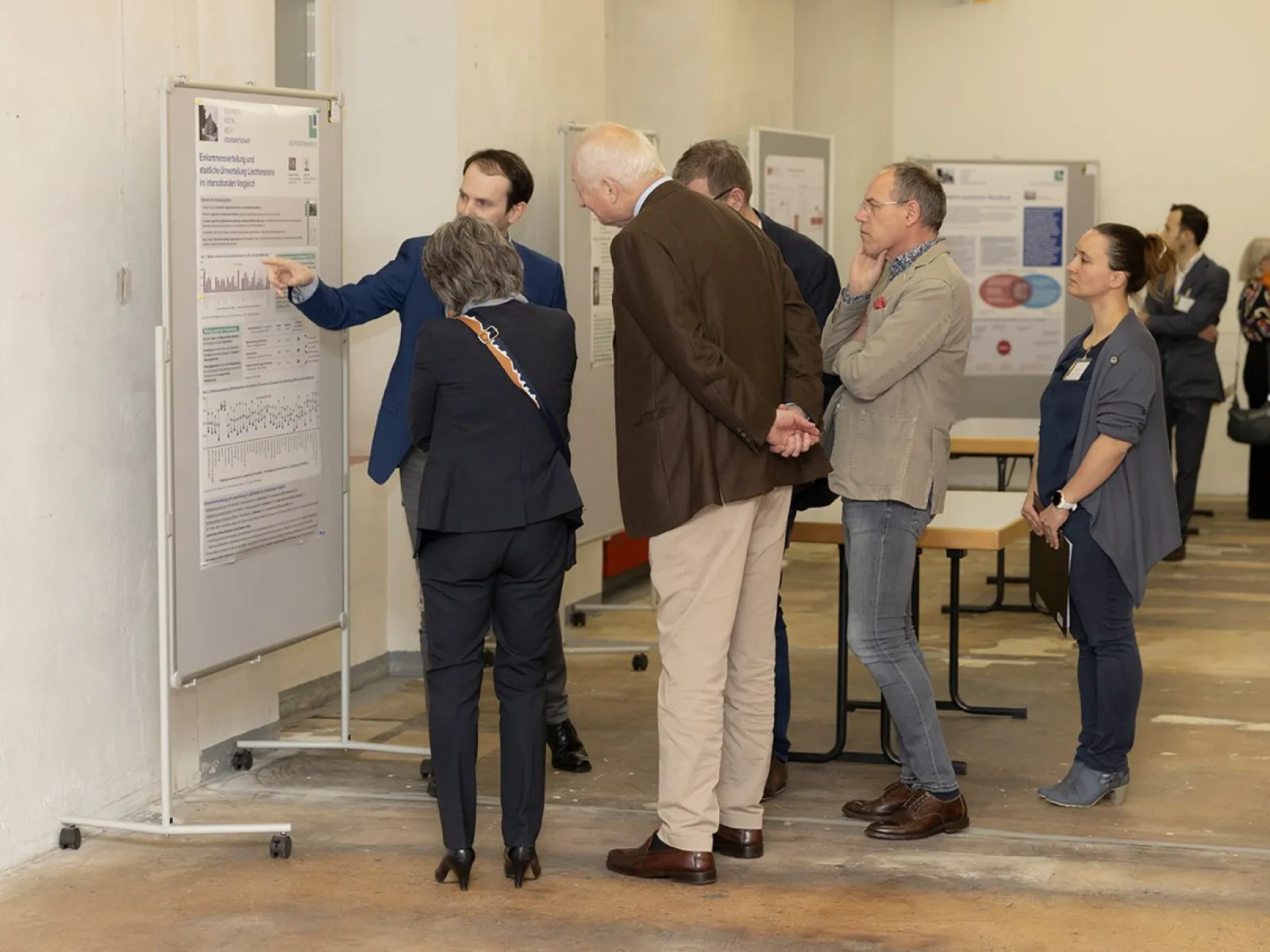 Eine Gruppe von Personen betrachtet eine wissenschaftliche Posterausstellung in einem Innenraum. Ein Mann zeigt auf ein Poster und erklärt den Inhalt. Im Hintergrund sind weitere Poster und Personen zu sehen.