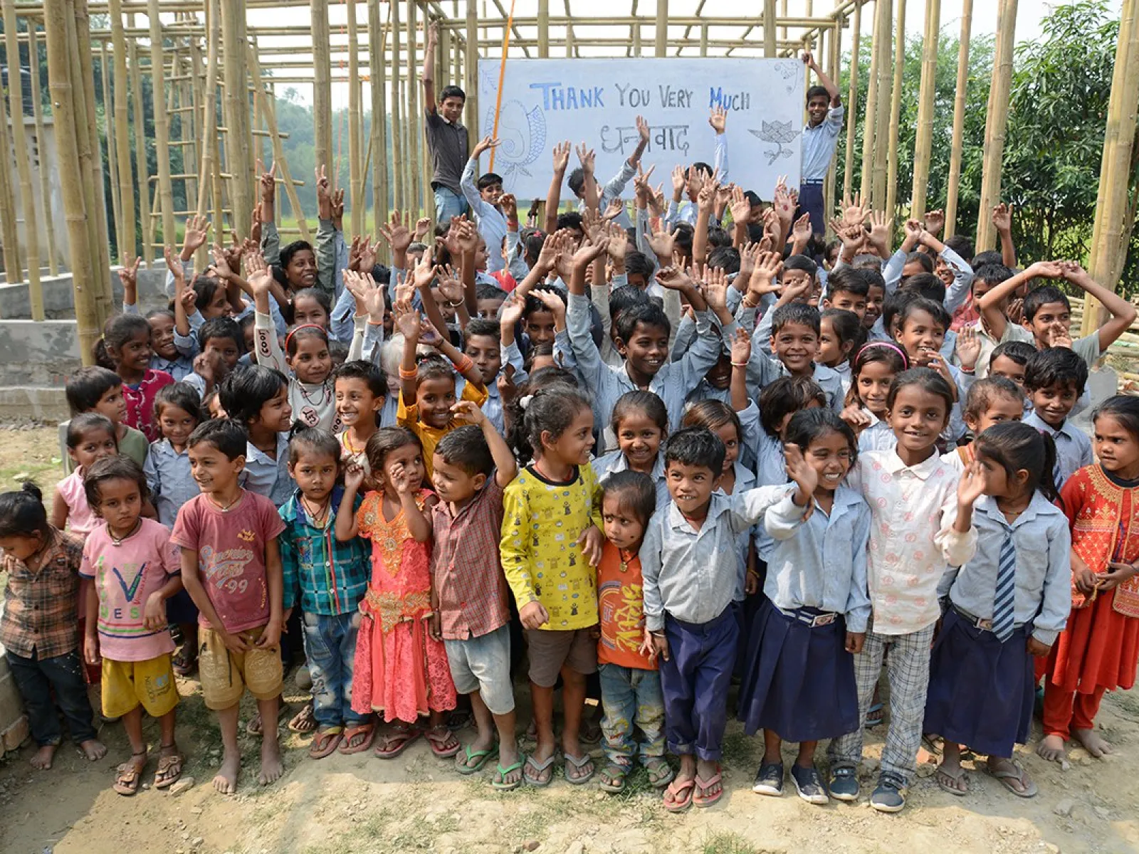 Eine große Gruppe fröhlicher Kinder steht vor einem im Bau befindlichen Schulgebäude in Bahuarwa, Indien. Im Hintergrund halten zwei Erwachsene ein Schild mit der Aufschrift "Thank You Very Much". Die Kinder lächeln und winken in die Kamera.