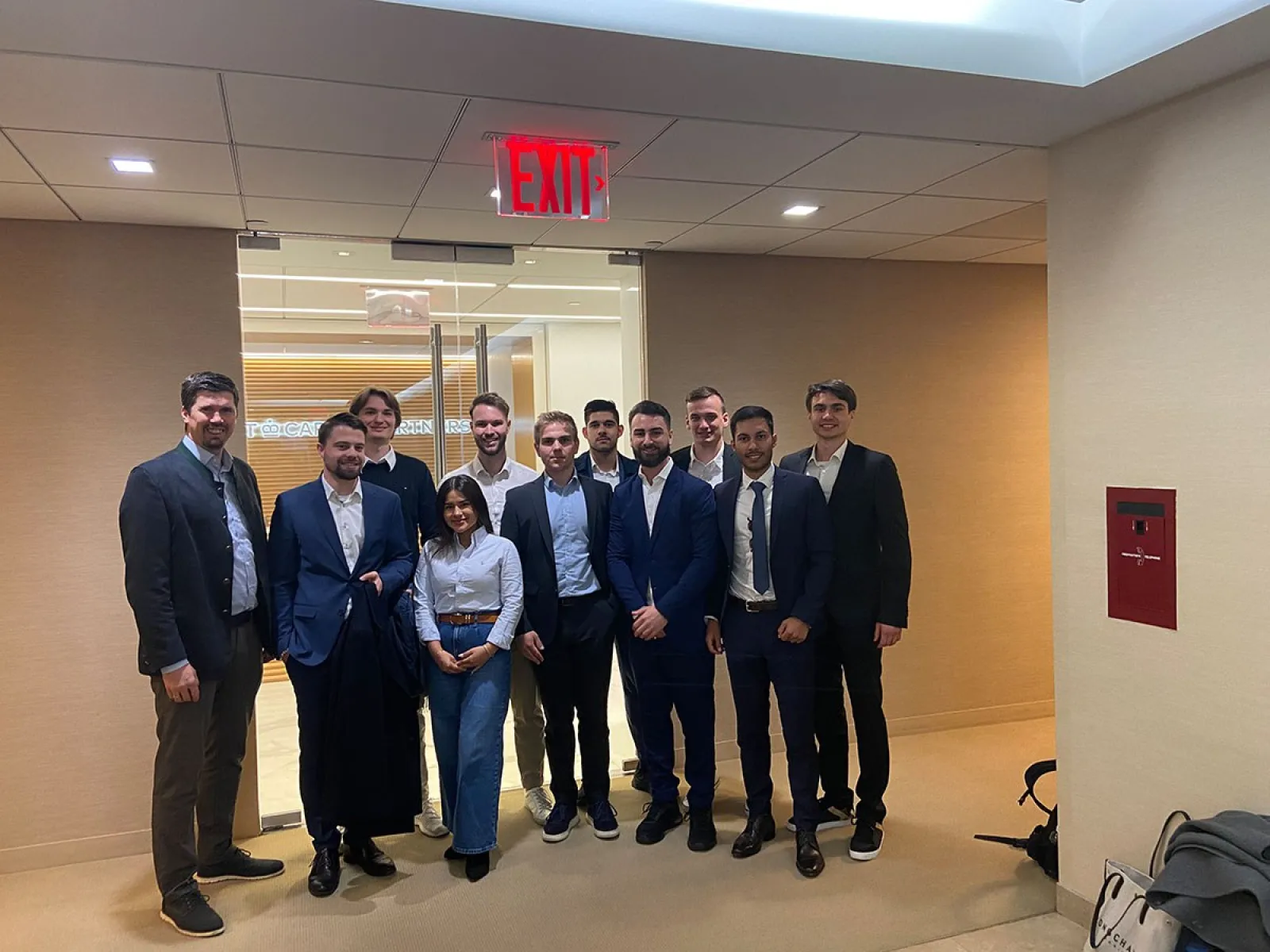 Eine Gruppe von Studierenden in Business-Kleidung steht in einem modernen Bürogebäude. Im Hintergrund ist eine Glastür zu sehen. Die Szene wirkt professionell und vermittelt den Eindruck einer Exkursion oder eines geschäftlichen Treffens.