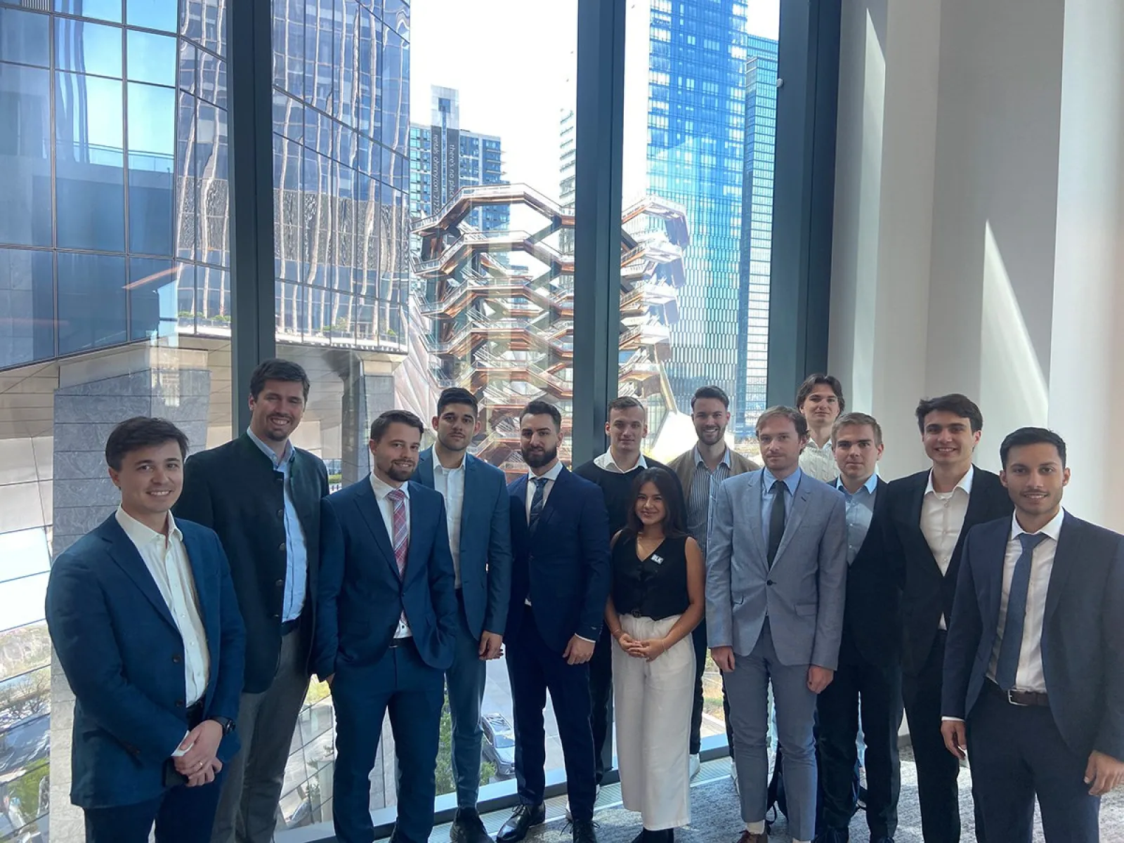 Eine Gruppe von Studierenden in Business-Kleidung steht vor großen Fenstern mit Blick auf moderne Architektur und Wolkenkratzer in New York City.