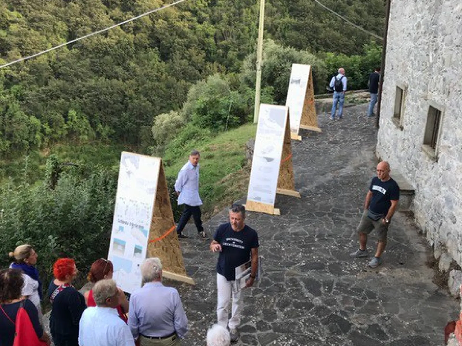 Eine Gruppe von Menschen betrachtet im Freien aufgestellte Tafeln mit Informationen. Die Szene spielt sich auf einem gepflasterten Weg neben einem alten Steingebäude ab, umgeben von üppiger, grüner Landschaft.