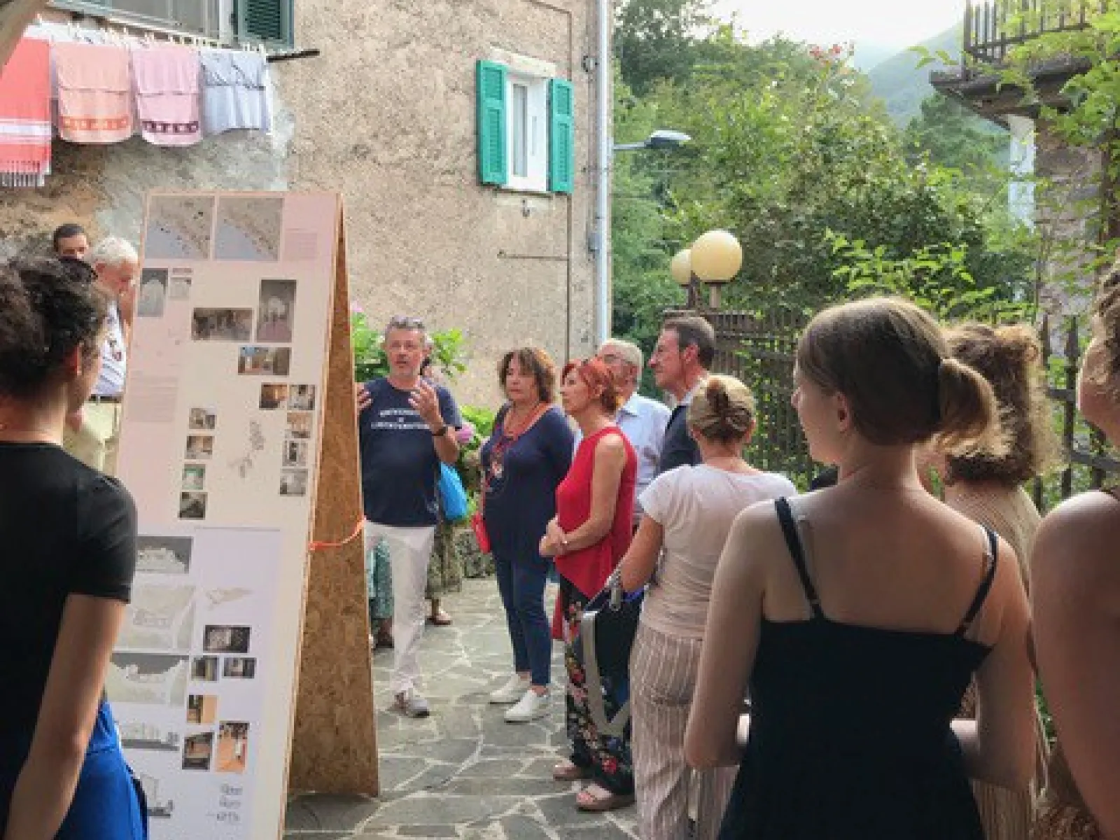Eine Gruppe von Menschen betrachtet eine Ausstellung im Freien in einem historischen Dorf. Im Vordergrund sind Informationstafeln zu sehen, im Hintergrund eine steinerne Hausfassade und grüne Hügel.