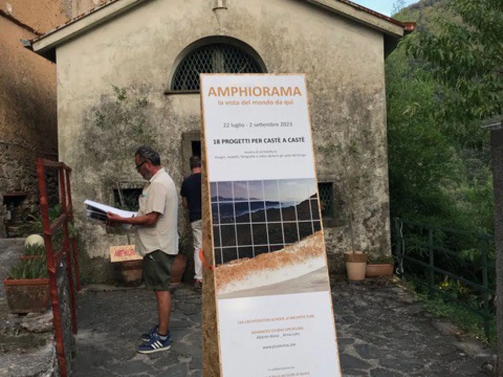 Ein Mann steht vor einem historischen Gebäude in Castè, Italien, und betrachtet ein Plakat mit der Aufschrift "Amphiorama". Das Plakat informiert über ein Projekt der Universität Liechtenstein im Rahmen des Advanced Studios Upcycling. Im Hintergrund sind die malerischen Hügel der Cinque Terre zu sehen.