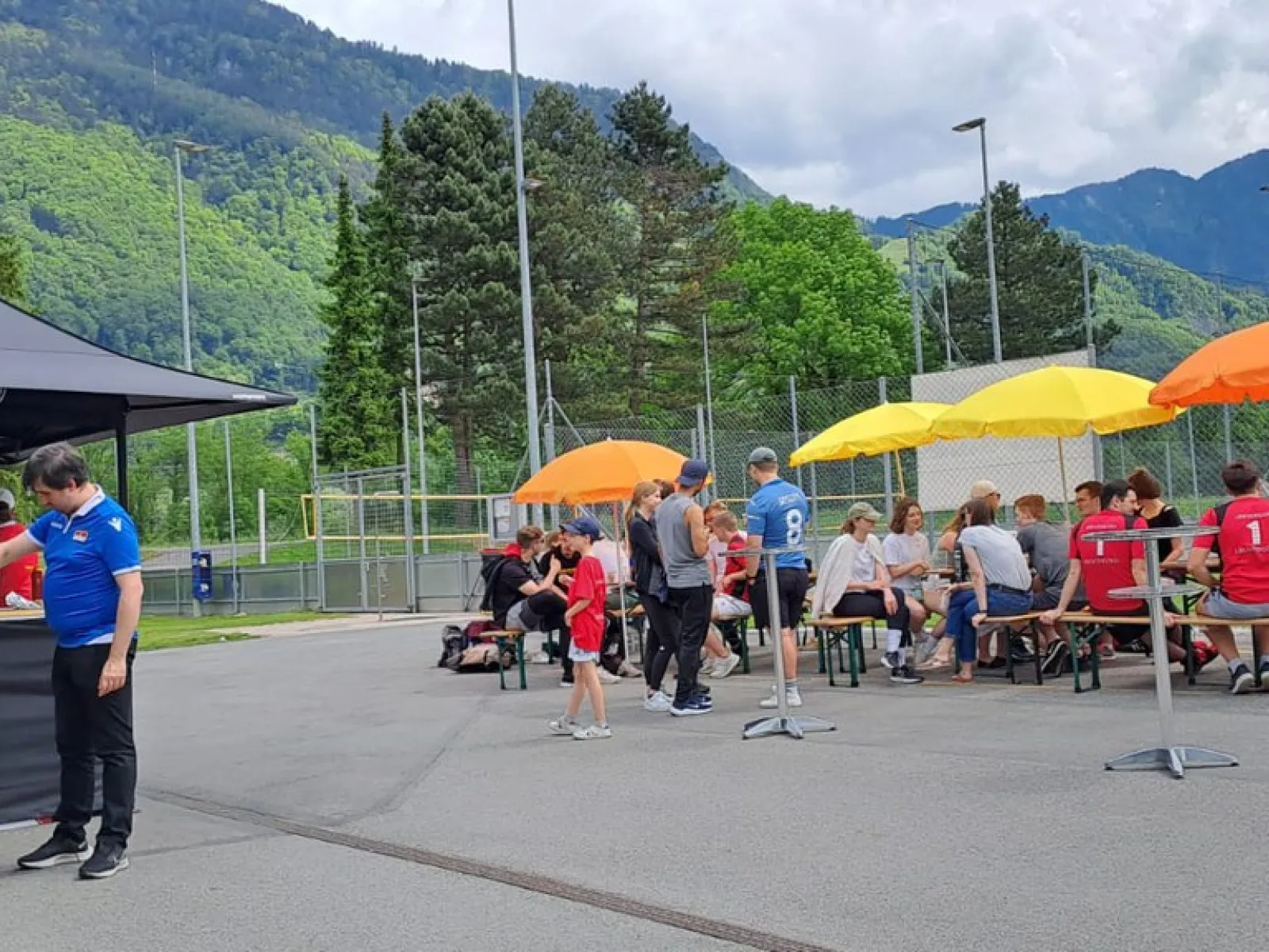 Teilnehmende des Völkerballturniers der Universität Liechtenstein sitzen unter bunten Sonnenschirmen auf Bänken. Im Hintergrund sind Berge und ein Zelt mit Verpflegungsstand zu sehen.