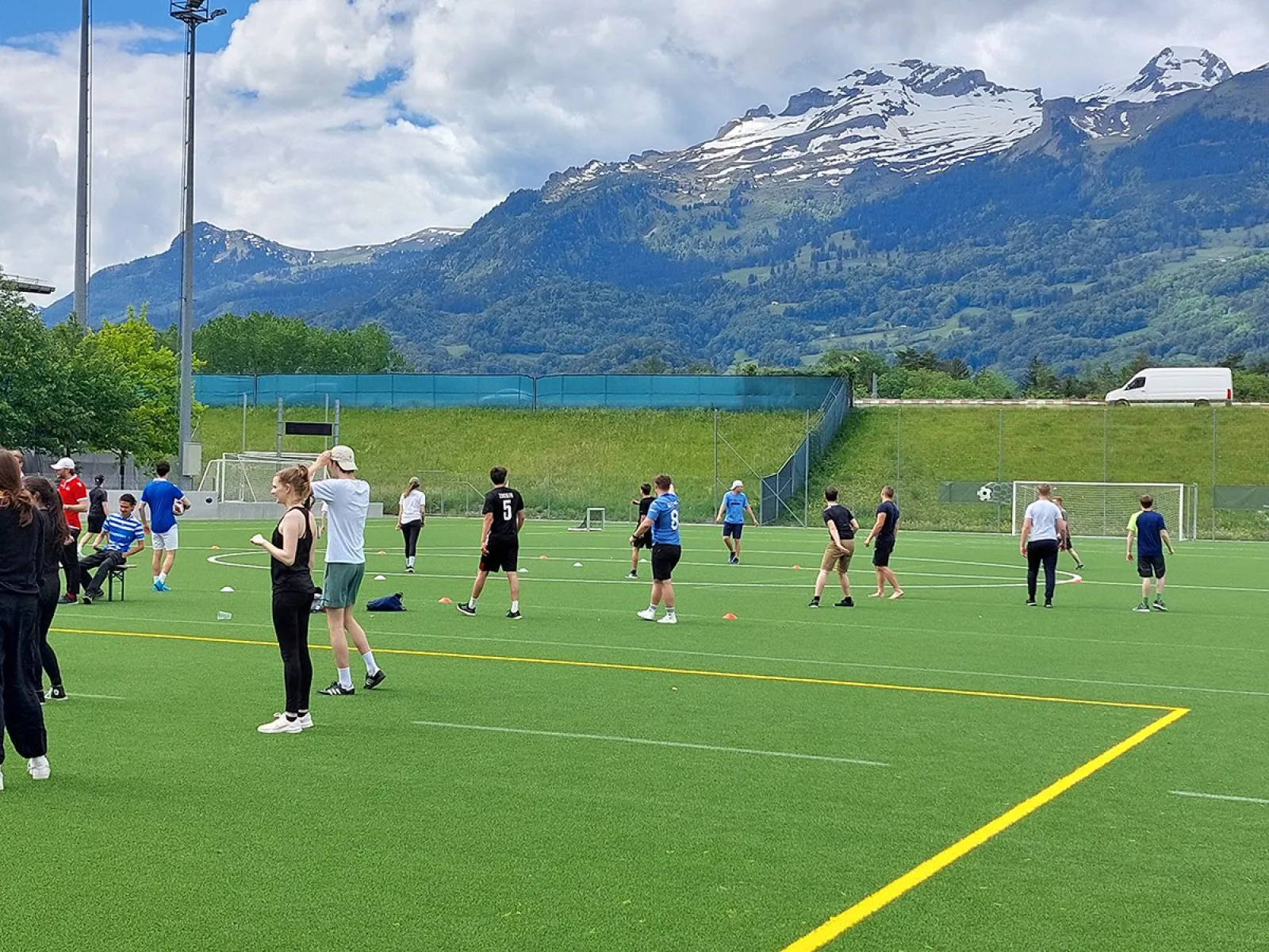 Eine Gruppe von Menschen spielt Völkerball auf einem grünen Sportplatz vor einer beeindruckenden Bergkulisse. Der Himmel ist teils bewölkt. Die Szene zeigt sportliche Aktivität und Gemeinschaftsgefühl bei sonnigem Wetter.