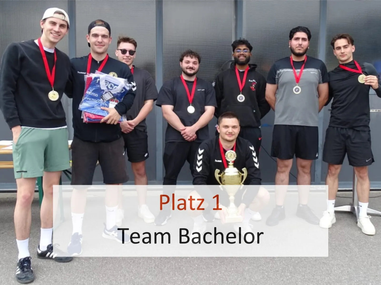Acht junge Männer stehen in sportlicher Kleidung vor einer Wand. Sie tragen Medaillen um den Hals. Einer kniet vorne und hält einen Pokal. Text im Bild: "Platz 1 Team Bachelor".