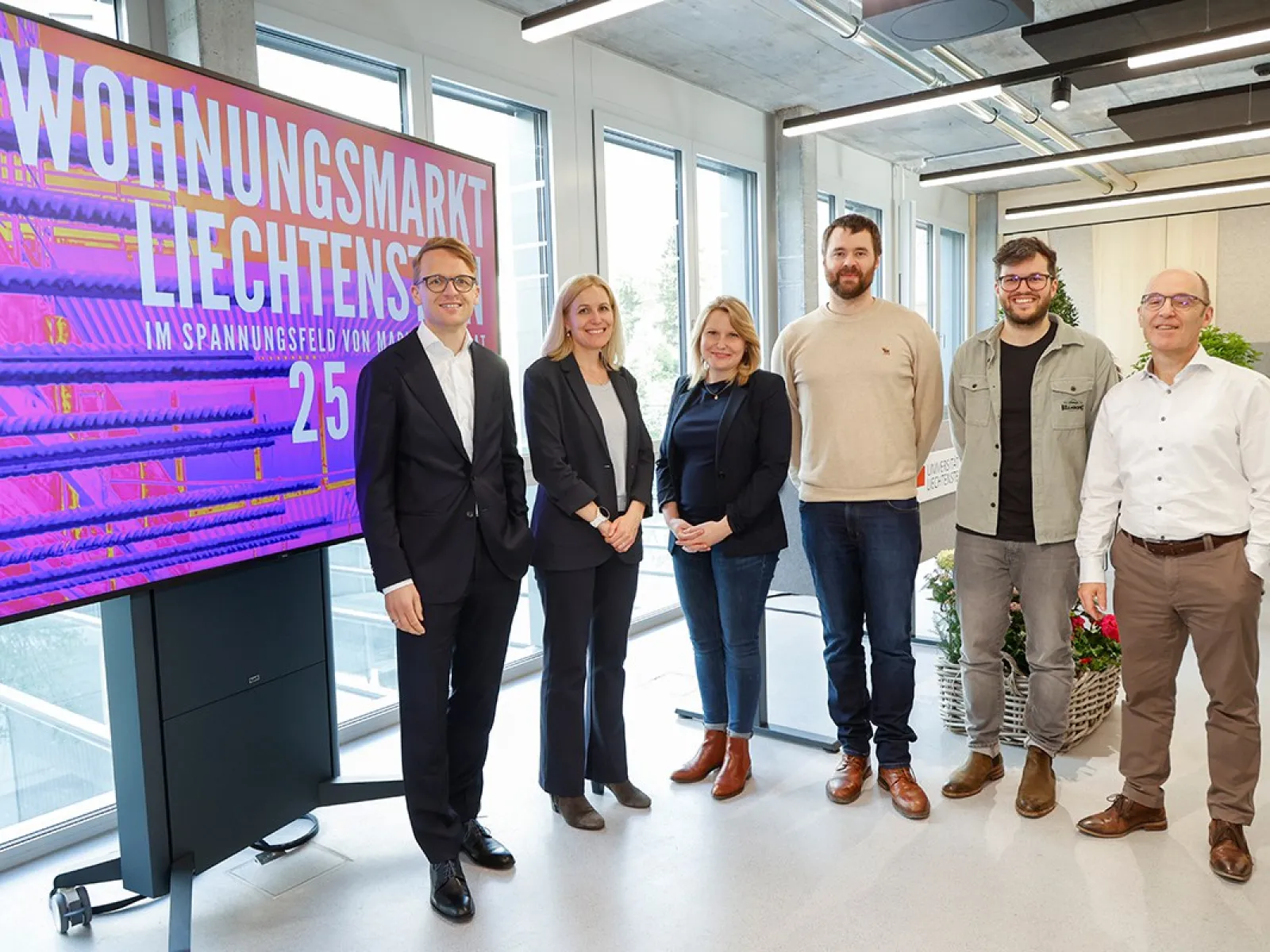 Gruppe von sechs Personen steht vor einem Bildschirm mit der Aufschrift "Wohnungsmarkt Liechtenstein". Die Personen lächeln und sind in einem modernen Raum mit großen Fenstern.