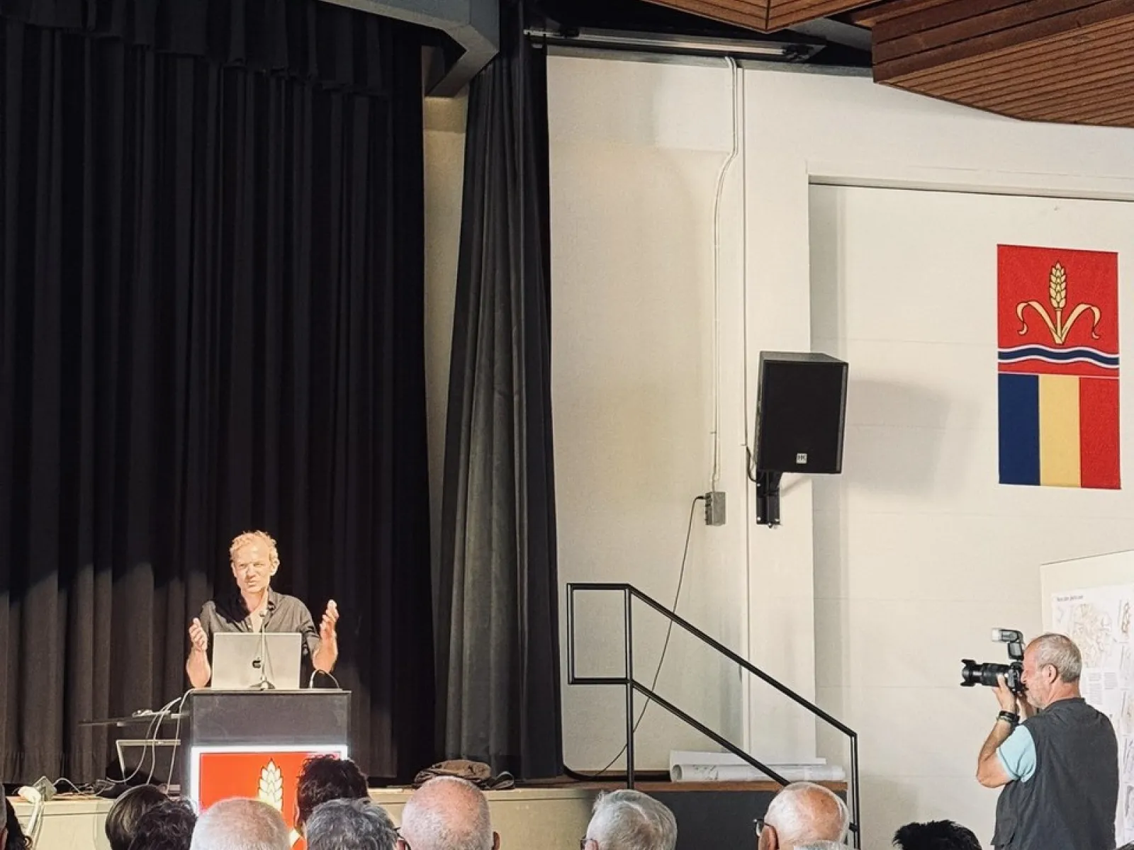 Ein Vortragender steht an einem Rednerpult vor einem Publikum in einem Saal. Im Hintergrund sind schwarze Vorhänge und ein Fotograf macht Bilder. An der Wand hängt ein Wappen.
