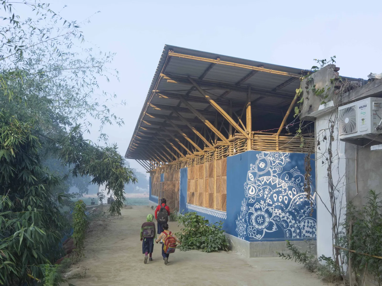 Kinder gehen auf einem sandigen Weg an einem blau bemalten Bambusgebäude mit weißem Ornamentmuster vorbei.