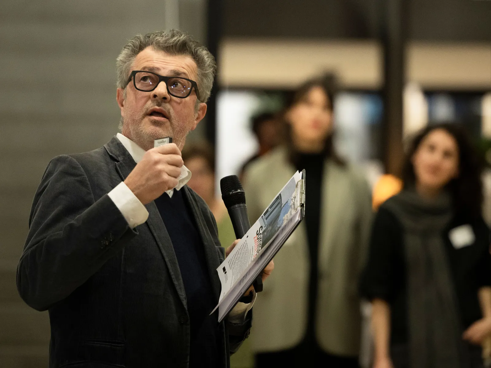 Ein Mann mit einer schwarzen Brille im Anzug spricht in ein Mikrofon