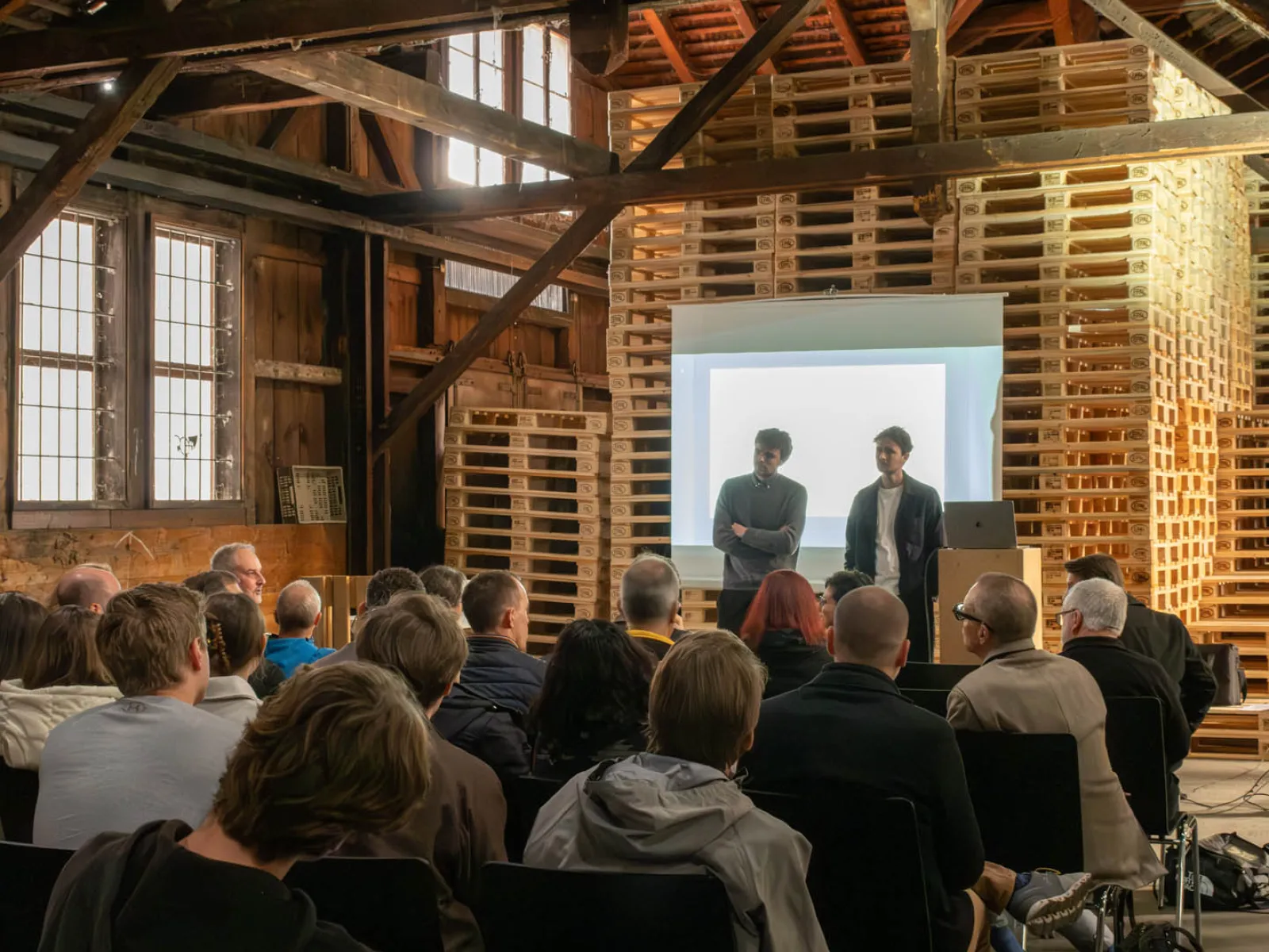 Zwei Männer stehen in einem Dachgeschoss udn präsentieren ein Projekt vor einem sitzenden Publikum