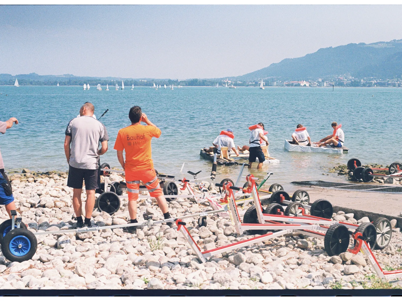 Mehrere Menschen am Ufer schauen auf selbstgebaute Boote in einem See. 