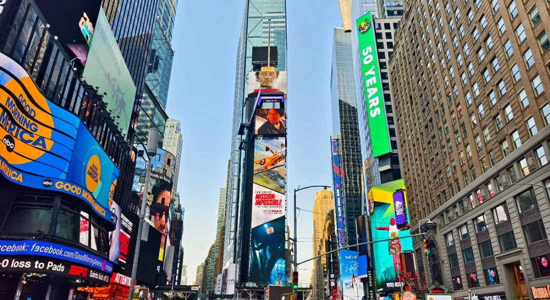 Blick auf den belebten Times Square in New York City mit hohen Gebäuden und leuchtenden Werbetafeln. Menschen und Autos beleben die Szene.