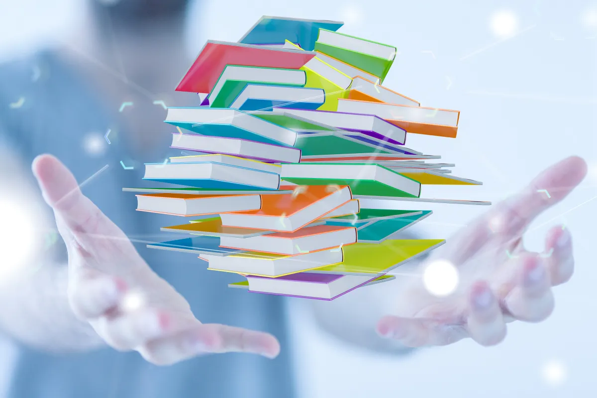 Schwebender Stapel bunter Bücher zwischen ausgestreckten Händen vor hellem Hintergrund, symbolisiert digitales Lernen.